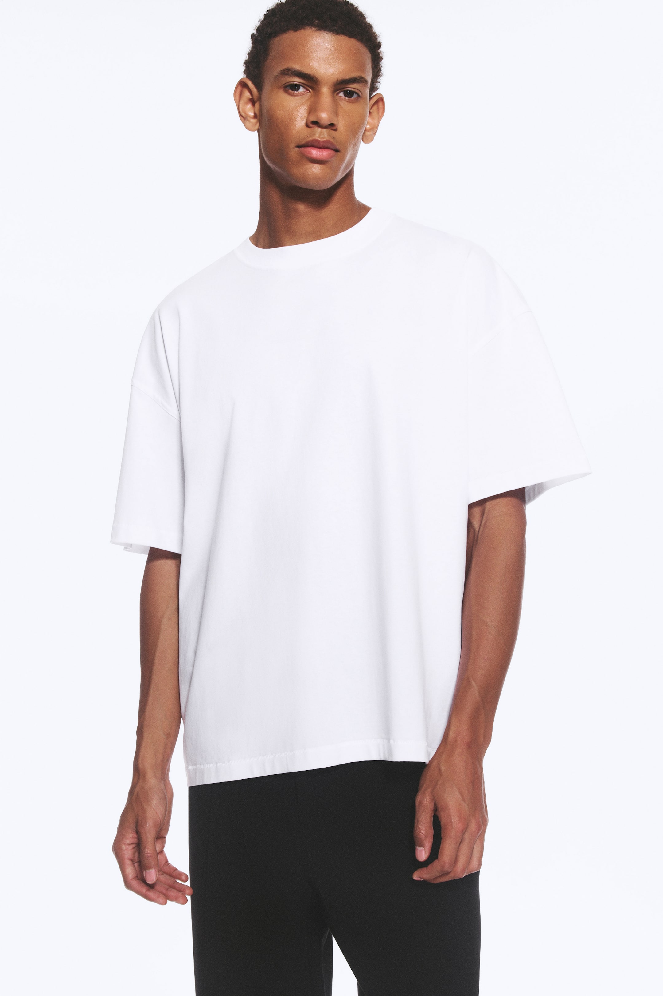 Agrandir l'image: Un t-shirt blanc oversize à coupe boxy est porté par le mannequin, avec un col rond, des épaules tombantes et des manches larges et amples. Le t-shirt est associé à un pantalon noir.