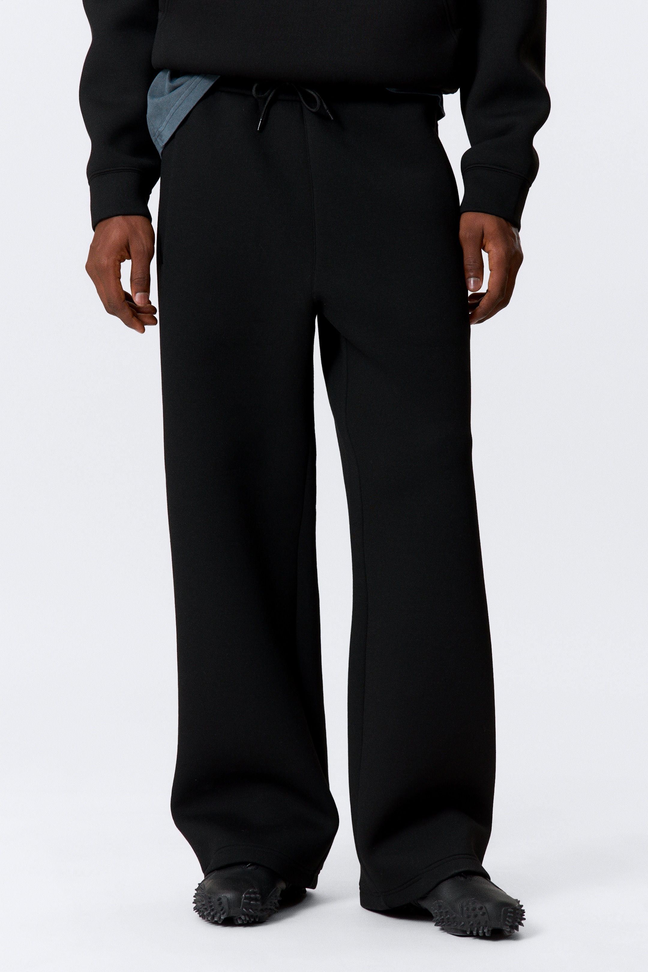 Agrandir l'image: Un pantalon noir à jambes larges est doté d'une taille élastiquée avec un cordon de serrage noir visible à l'avant. Le pantalon tombe dans une coupe ample, s'étendant jusqu'au sol et s'accumule légèrement sur les chaussures noires du porteur.