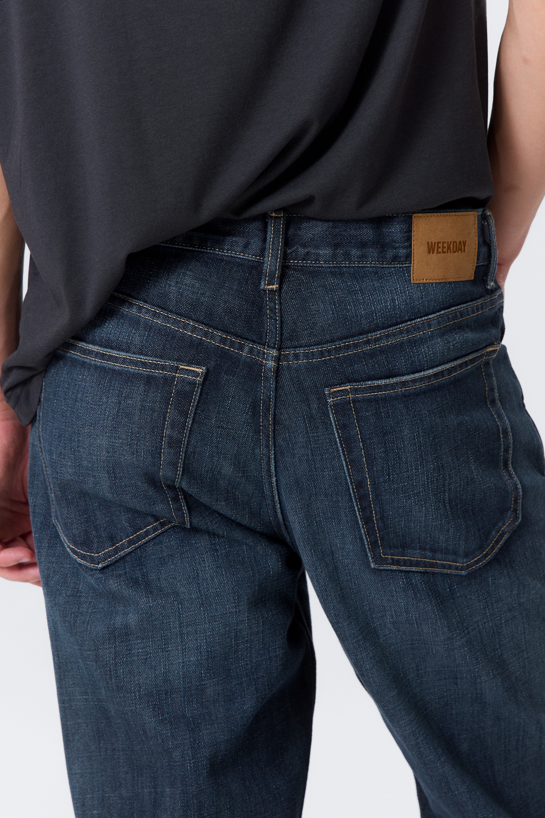 View larger image: Dark wash denim jeans are being worn, seen from the back, slightly angled to the right. They are featuring two back patch pockets with prominent light brown topstitching and a rectangular light brown "WEEKDAY" branded patch on the right side of the waistband, displaying a relaxed fit.