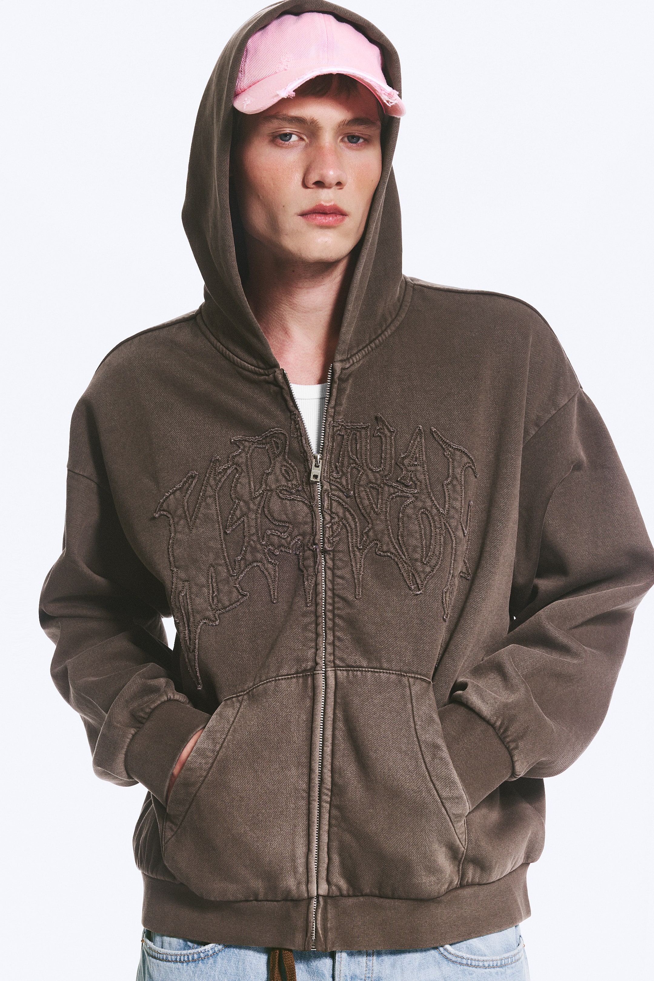 Visualizza immagine più grande: Una felpa con cappuccio marrone con zip è presentata frontalmente, caratterizzata da un aspetto vissuto e lavato e da un intricato ricamo marrone scuro che ricorda un testo stilizzato sul petto. Presenta una zip color argento lungo la parte anteriore, un cappuccio attaccato, polsini a coste, un orlo a coste e due tasche frontali a marsupio, con una maglietta bianca visibile sotto la zip parzialmente aperta.