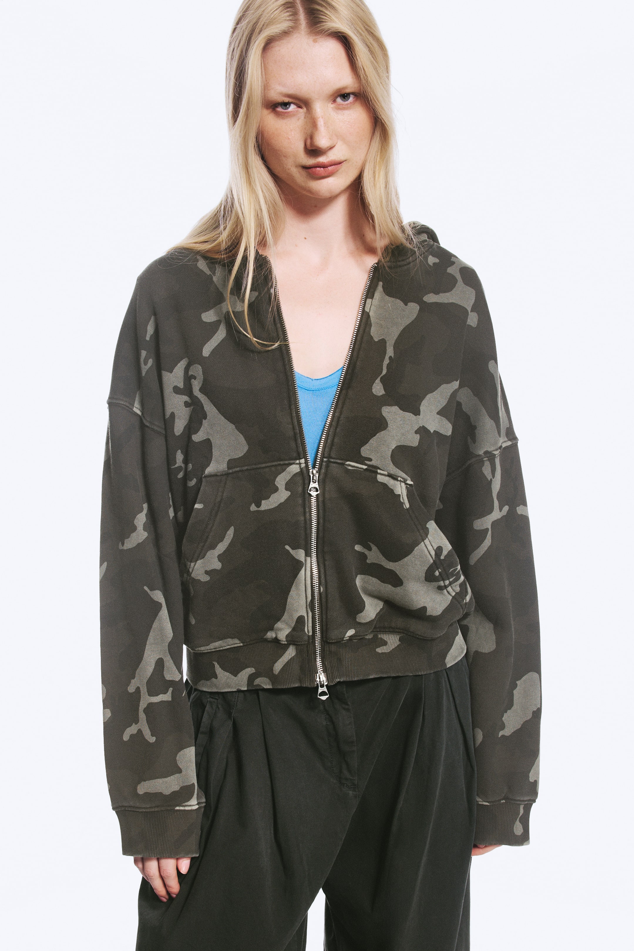 Visualizza immagine più grande: Una felpa oversize con cappuccio e zip, con stampa camouflage verde scuro, indossata dalla modella, abbinata parzialmente aperta sopra un top blu. La felpa presenta spalle scese, polsini a costine e tasche a marsupio divise, ed è abbinata a pantaloni scuri.