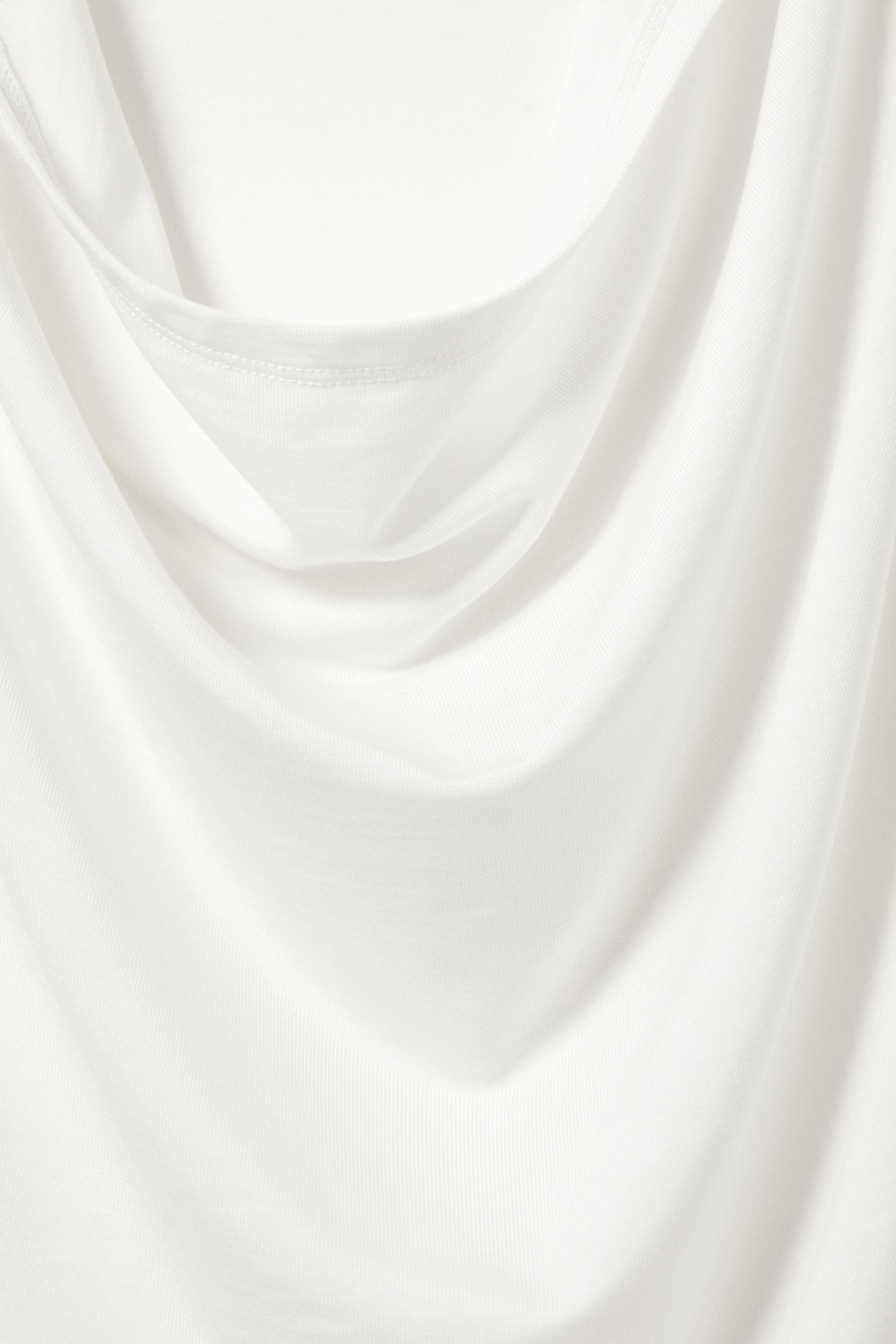 Se større billede: Et hvidt strikket stof bliver draperet, hvilket skaber bløde folder og kurver. En færdig kant med synlige syninger danner en cowl-neck-stil detalje øverst.