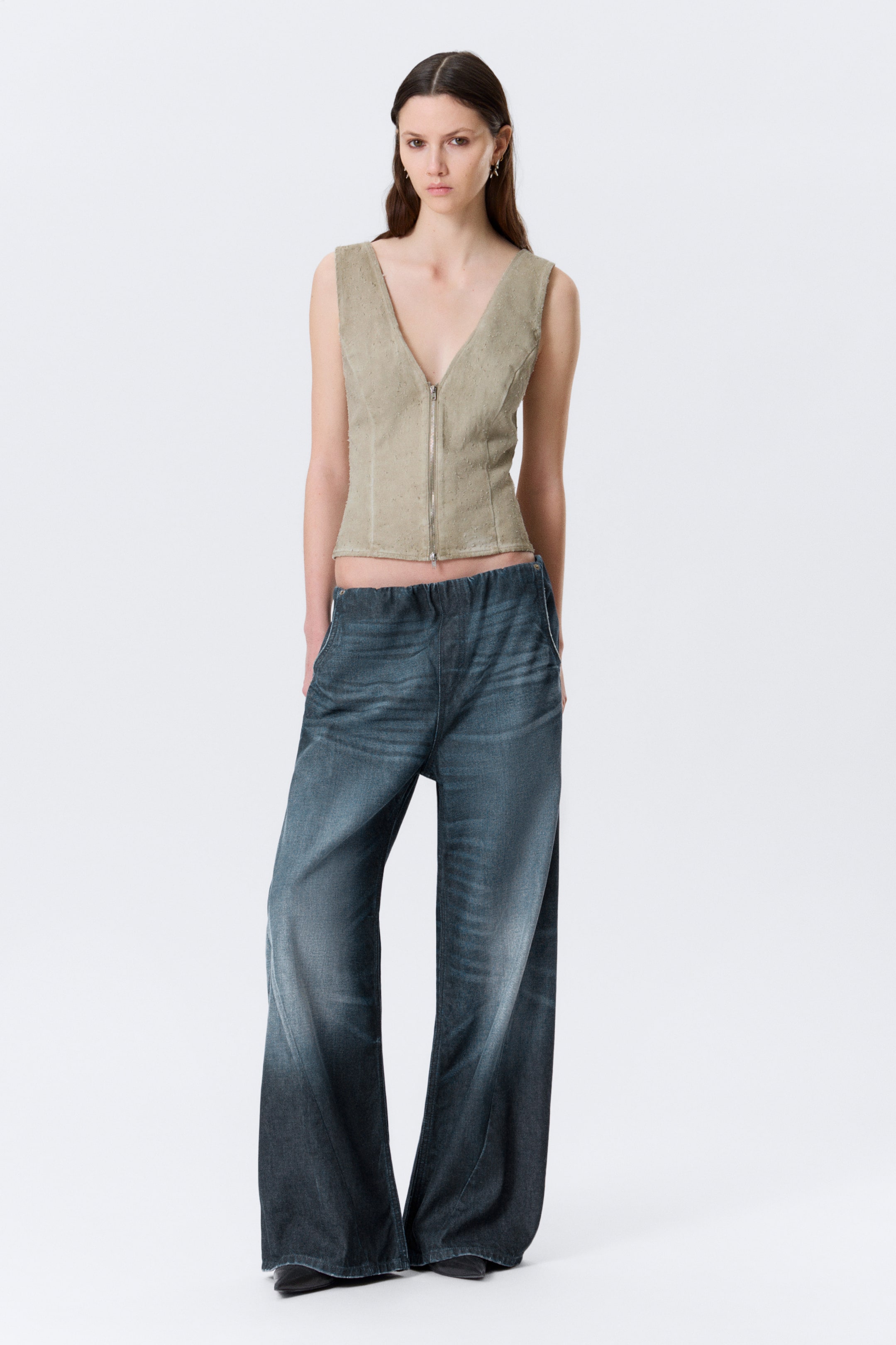 Grotere afbeelding bekijken: Laagzittende jogger-stijl jeans worden gedragen door het model. Deze ontspannen, loszittende jeans, gemaakt van stug denim, hebben een elastische tailleband en schuine zijzakken waarin het model haar handen rust. Een nep gulpstiksel is zichtbaar aan de voorkant en de jeans vertoont een donkere wassing die naar de onderkant vervaagt.