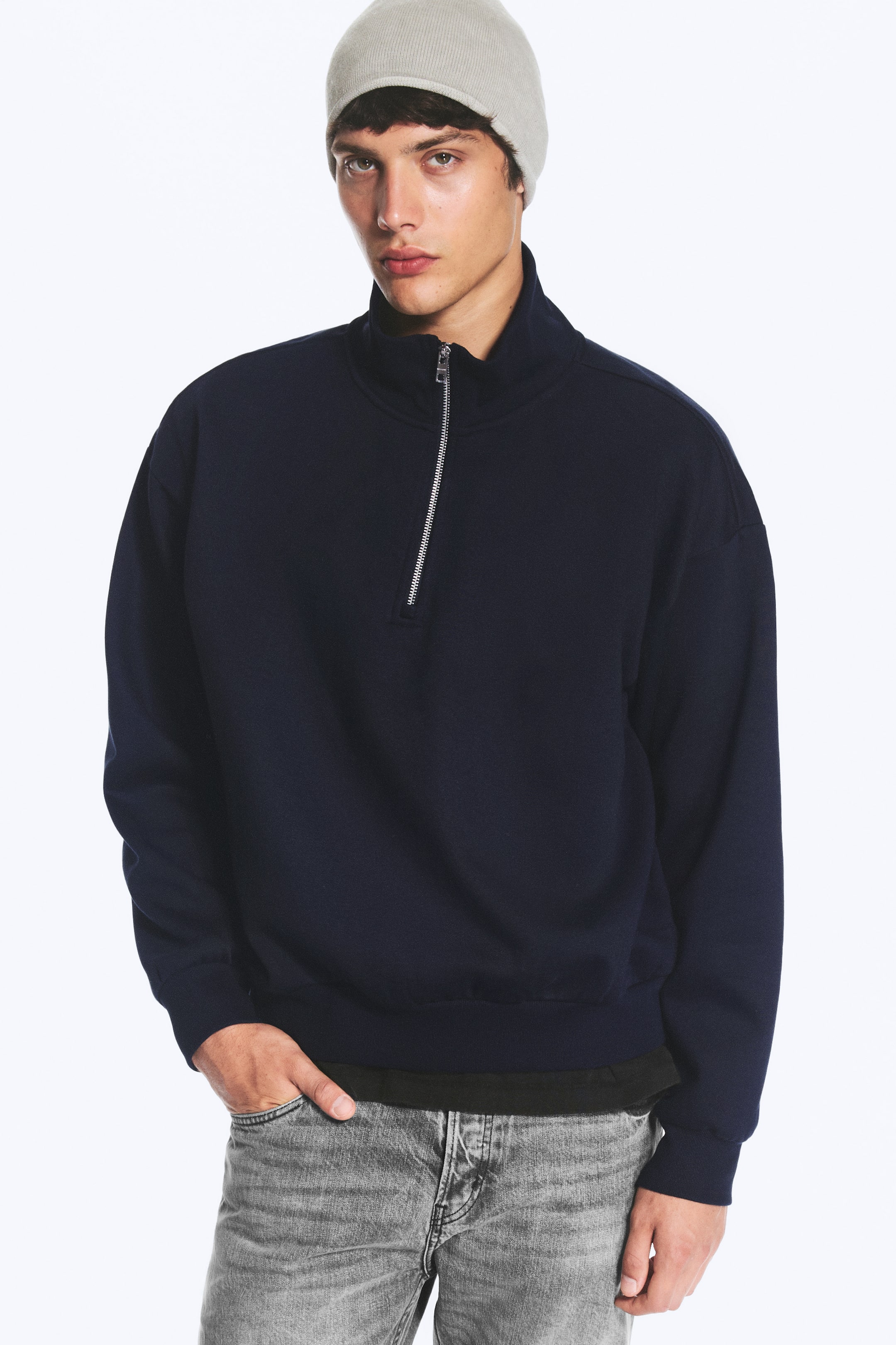 View larger image: The model is wearing a navy blue half-zip sweatshirt featuring a high ribbed collar, a silver metal zip and dropped shoulders. The sweatshirt is styled with grey jeans and a light grey beanie.