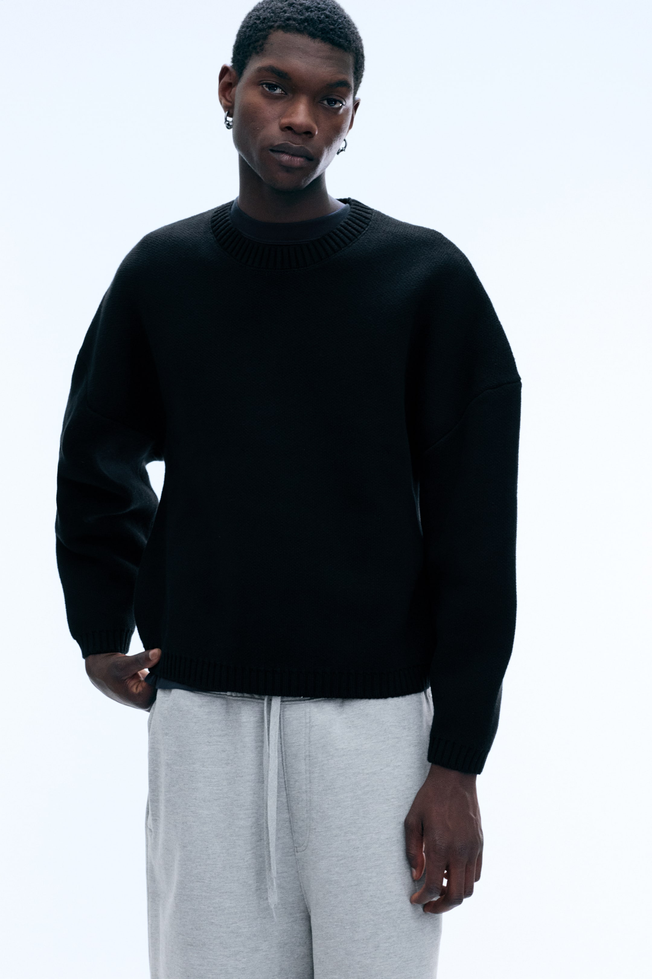 Agrandir l'image: Un pull noir oversize à col rond, doté d’épaules tombantes et de finitions côtelées, est porté par le mannequin. Le pull est associé à un t-shirt sombre à col rond et à un pantalon gris clair à cordon de serrage.