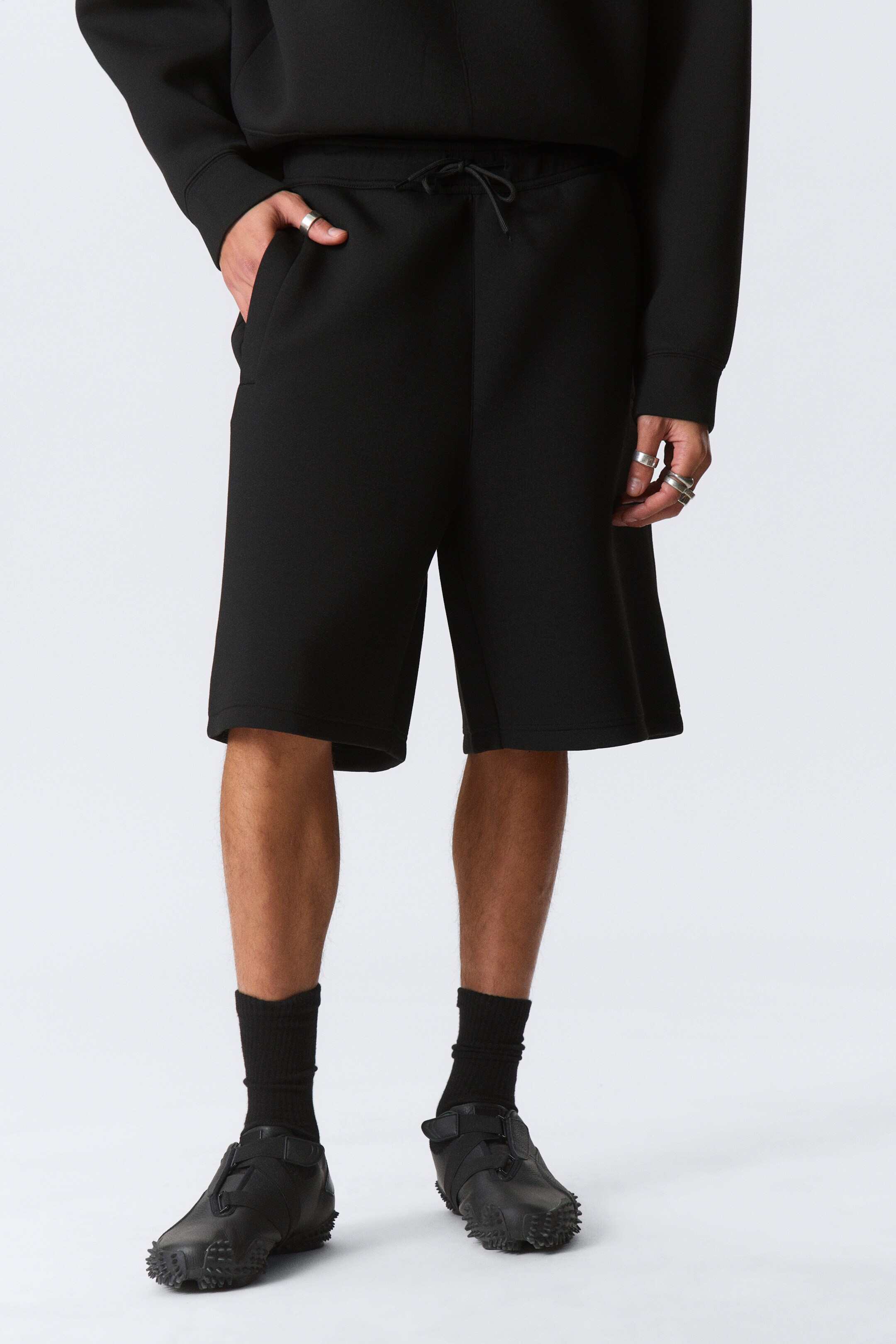 Vis større bilde: Svarte shorts har en elastisk linning med en synlig snor, og en hånd hviler i en sidelomme. De avslappede shortsene strekker seg til rett over knærne.