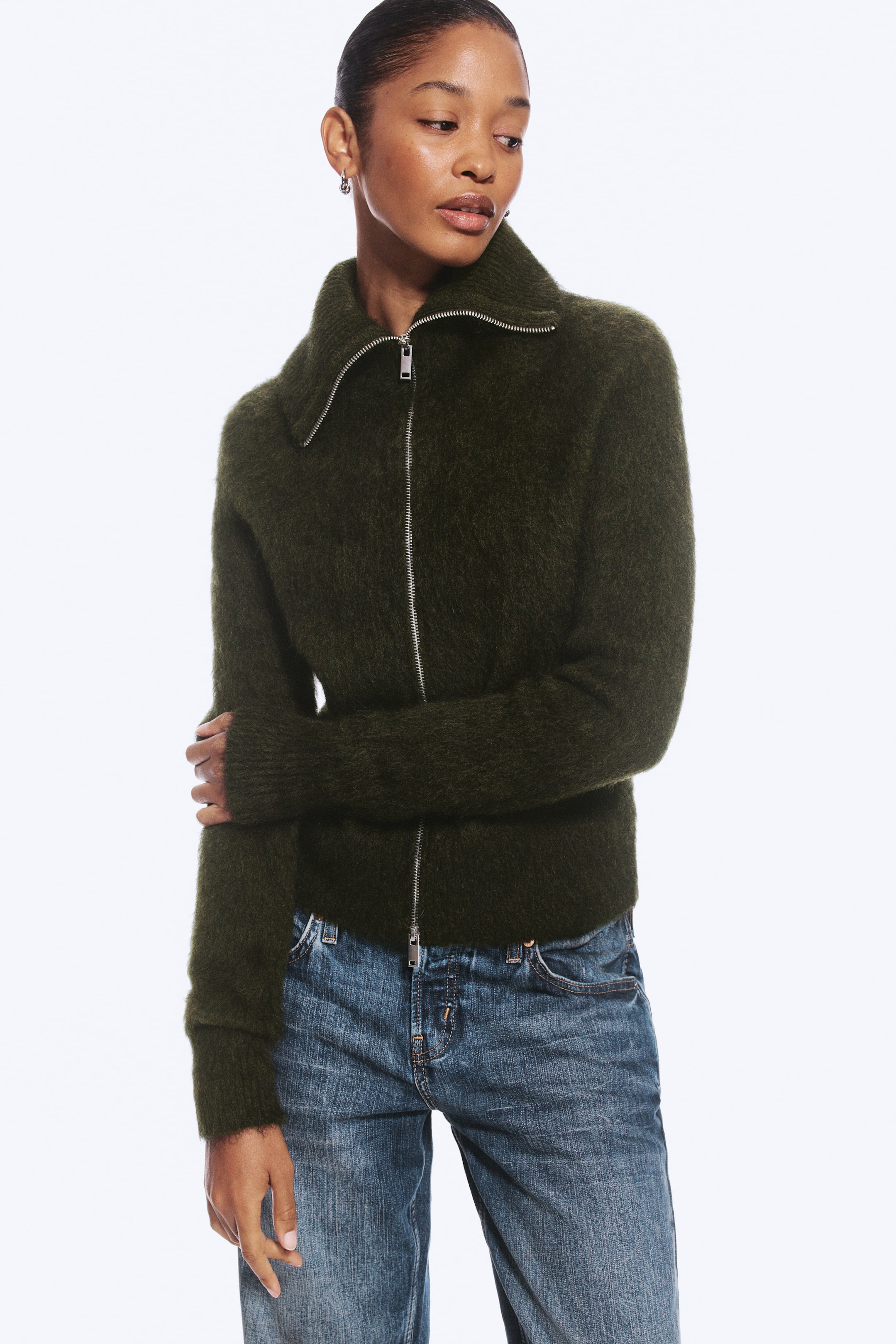 Visualizza l'immagine ingrandita: Una cardigan in maglia pelosa di colore verde scuro, con colletto alto e zip frontale argentata a doppio cursore, indossata dalla modella. La modella abbina la cardigan a jeans blu, tenendo in mano una delle lunghe maniche a coste.
