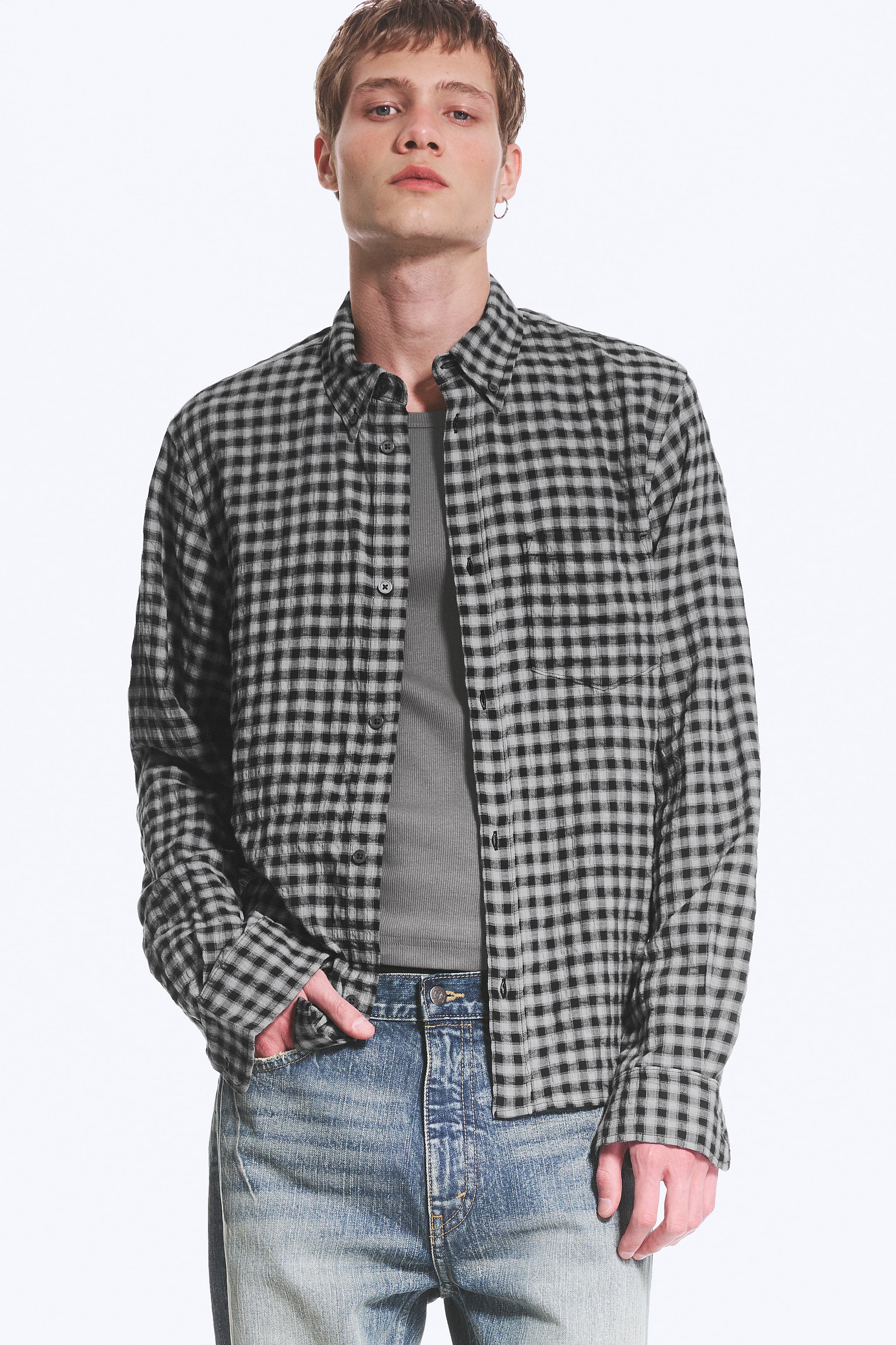 Agrandir l'image: Une chemise à carreaux vichy noire et gris clair pour homme est présentée de face, avec une texture légèrement froissée, un col boutonné et une poche plaquée sur la poitrine à gauche. Elle est portée ouverte sur un t-shirt gris côtelé, avec ses manches longues se terminant par des poignets boutonnés.