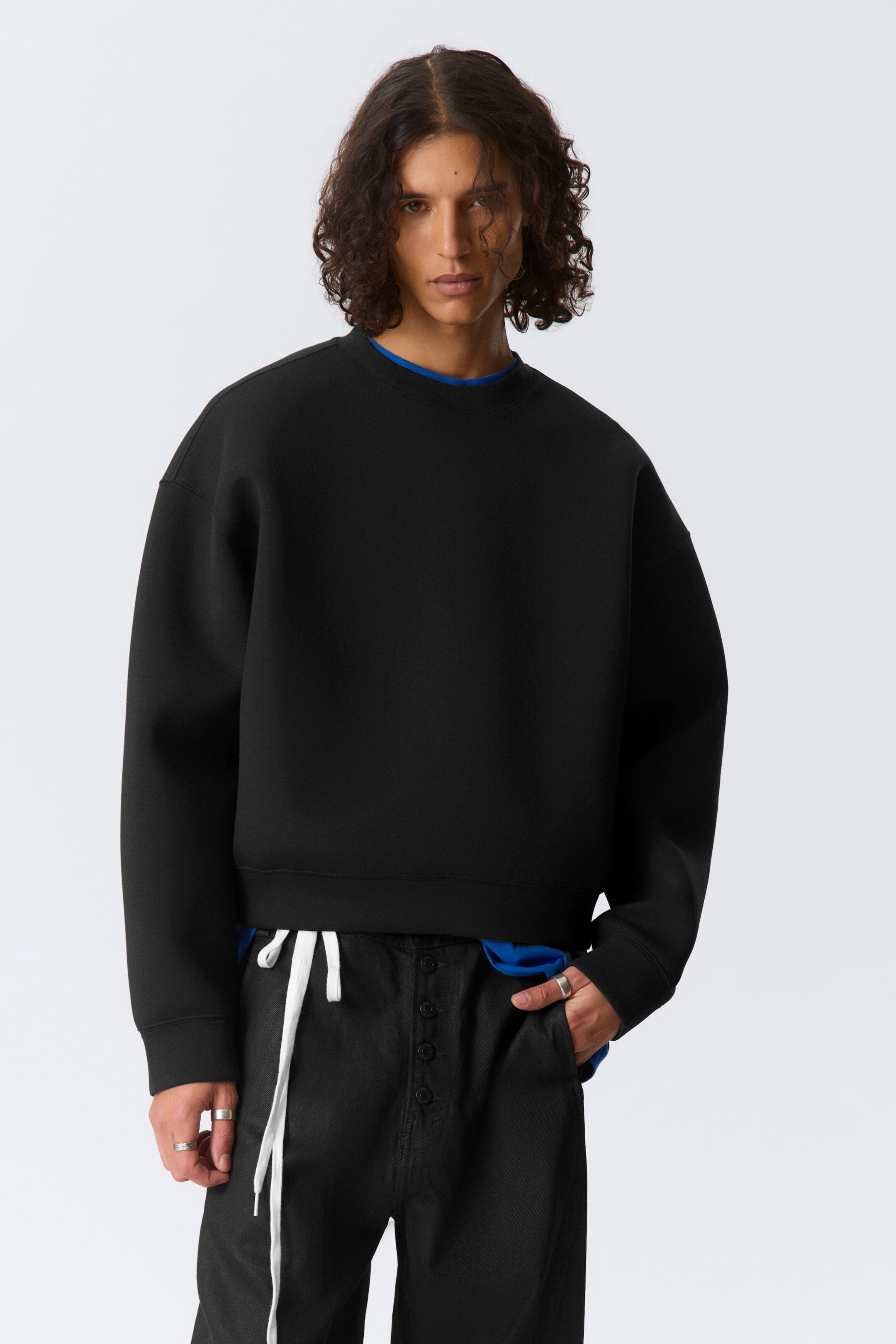 Se større billede: En sort crew neck sweatshirt præsenteres forfra, med en oversized, struktureret pasform med dropped skuldre, ribbede manchetter og en cropped kant. En blå crew neck skjorte er synlig nedenunder, og titter frem ved kraven og forneden.