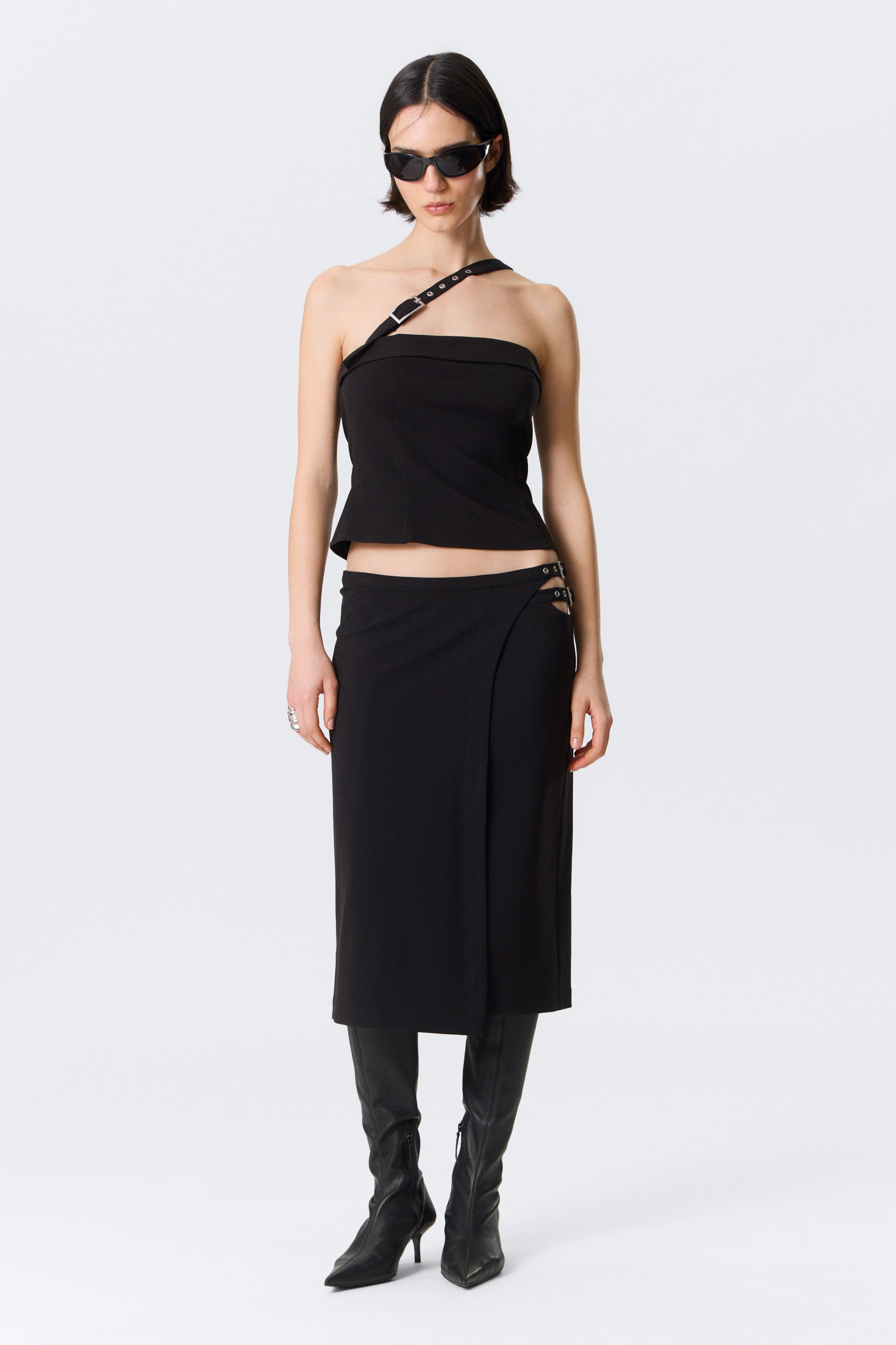View larger image: A black strapless top featuring a single buckled strap extending over the left shoulder and a structured bandeau neckline. It is styled with a black midi skirt, designed with a wrap-style front and metal ring details on the right hip.