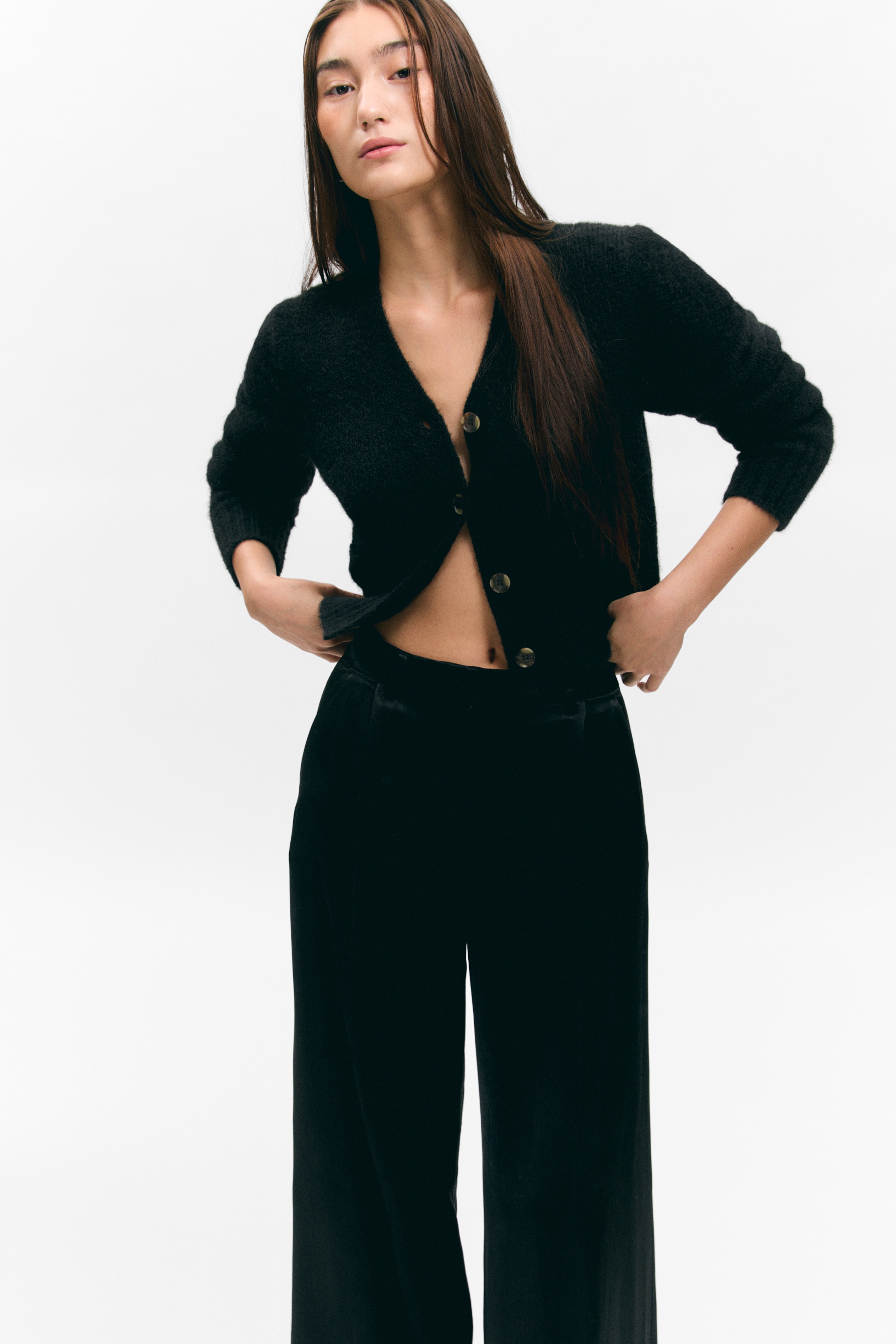 Agrandir l'image: Un cardigan en tricot noir et duveteux est présenté de face, avec un col en V et quatre boutons de couleur claire, certains déboutonnés pour mettre en valeur sa coupe courte et décontractée. Il est porté avec un pantalon noir à jambes larges qui semble taille haute.