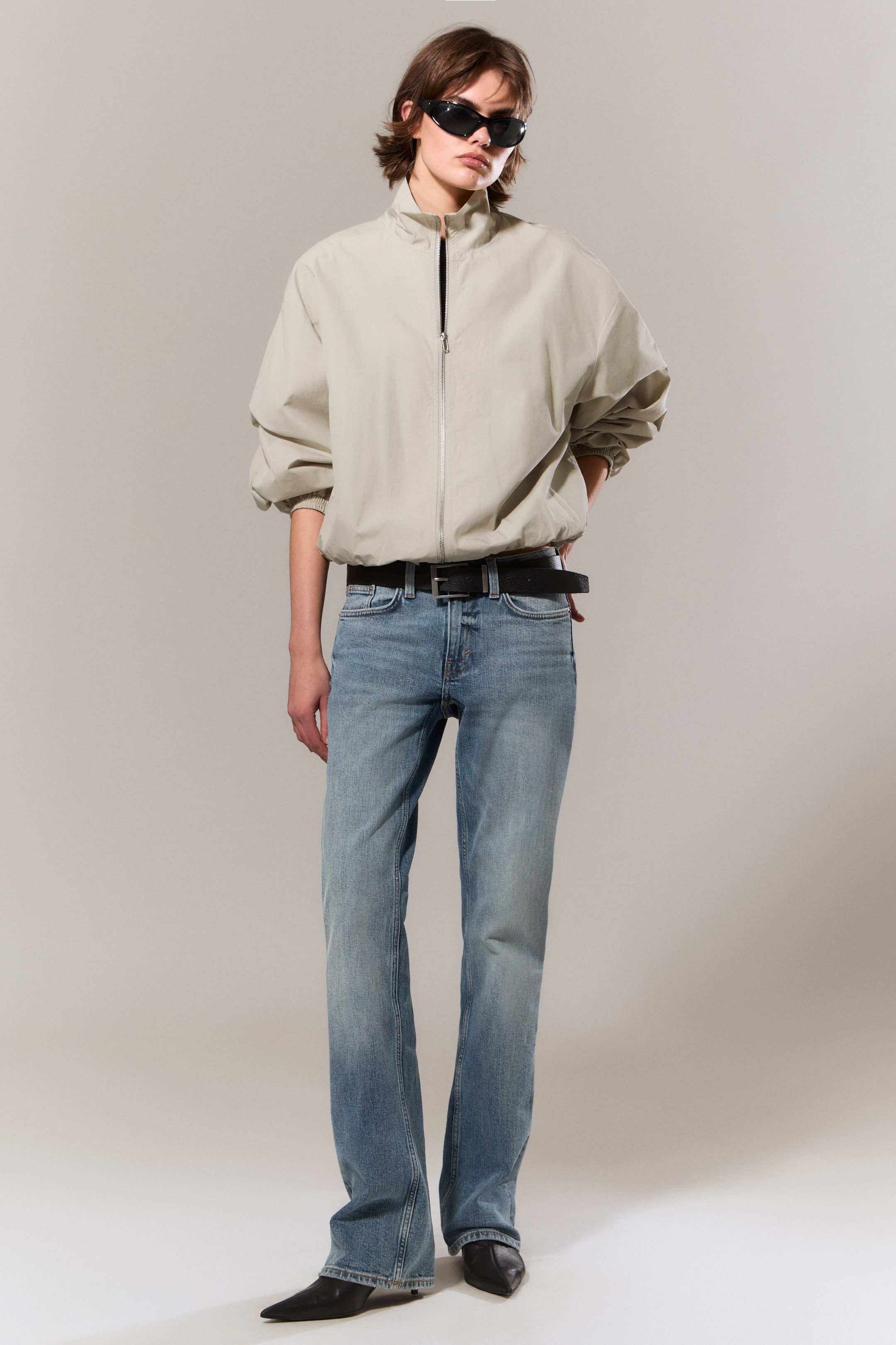 Größeres Bild anzeigen: Eine hellbeige Bomberjacke wird aus einem leichten Winkel präsentiert und zeigt einen Stehkragen sowie einen teilweise geöffneten Reißverschluss, der ein dunkles Innenfutter enthüllt. Sie verfügt über voluminöse Ärmel mit elastischen Bündchen und einem elastischen Saum, was eine entspannte, blousonartige Passform schafft.