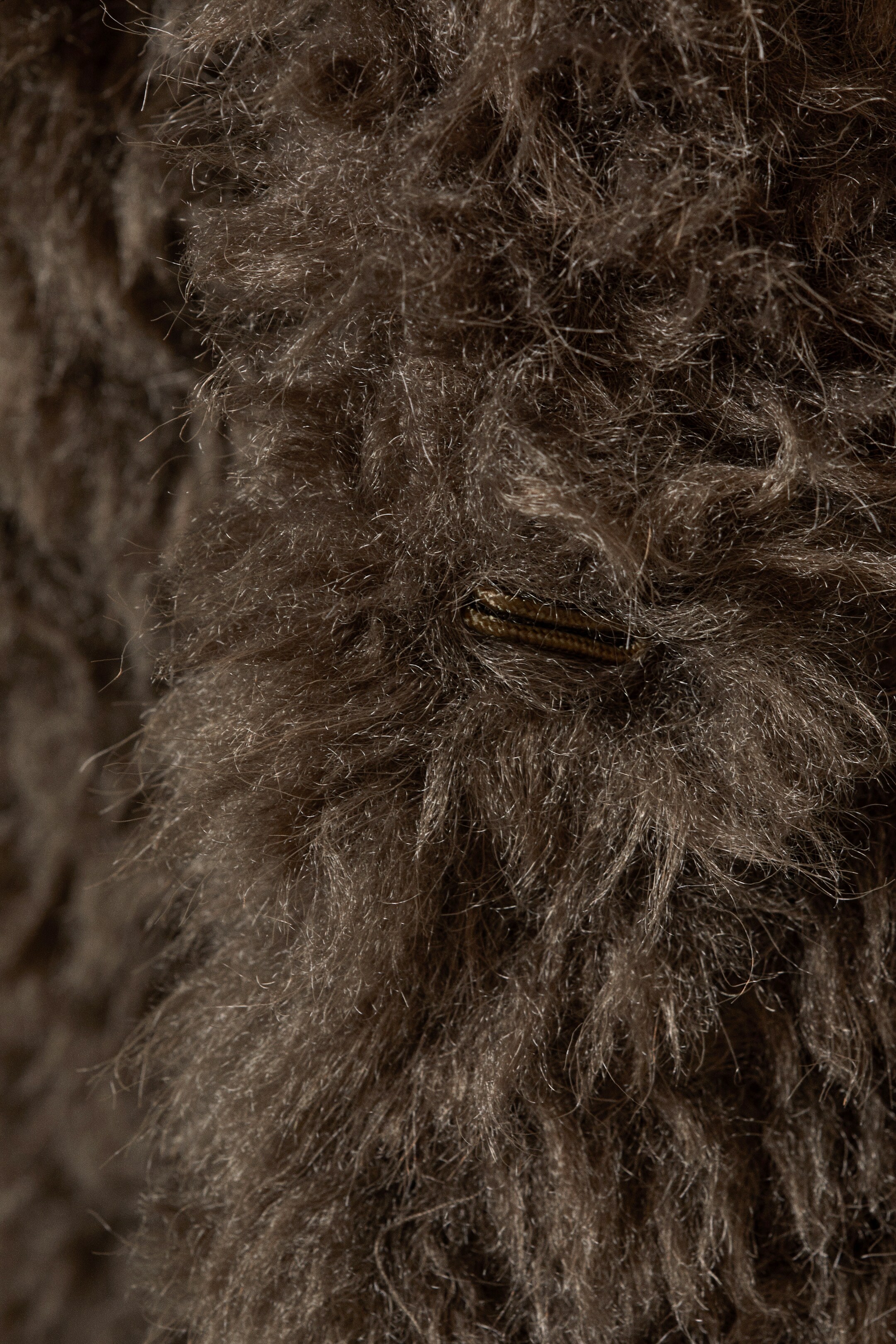 Agrandir l'image: Le produit présente un matériau brun foncé et hirsute avec de longues fibres texturées. Une boucle ou boutonnière sombre, orientée horizontalement, est partiellement visible, intégrée dans la texture dense.