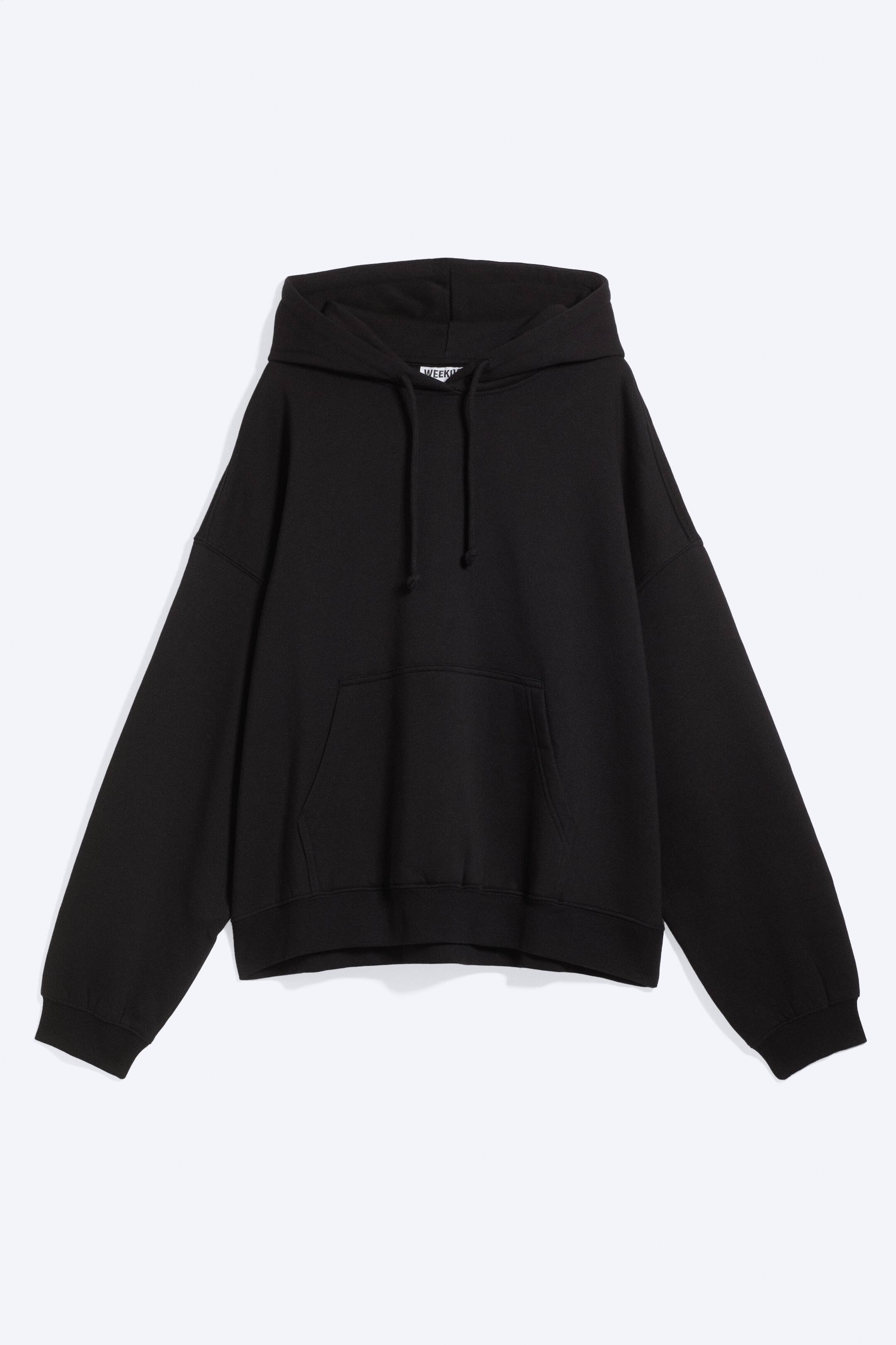 Ver imagen más grande: Una sudadera con capucha negra está orientada de frente y presenta un corte oversize. Muestra una capucha con cordones negros, un gran bolsillo tipo canguro en la parte frontal y puños y bajo de canalé.