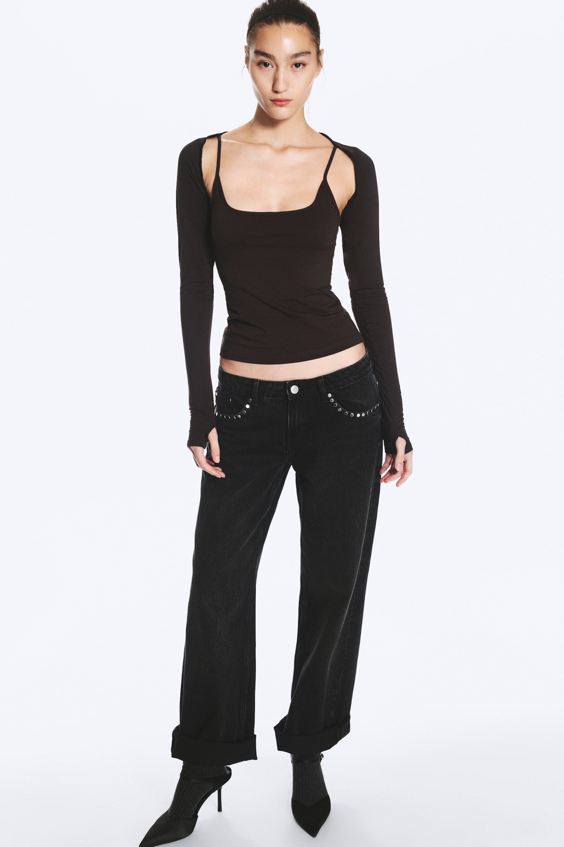 Visualizza immagine più grande: Un bolero nero slim fit con apertura frontale e maniche lunghe con fori per i pollici viene indossato dalla modella. Il bolero è sovrapposto a una canotta nera coordinata con scollo tondo, mettendo in risalto il design con spalle scoperte.