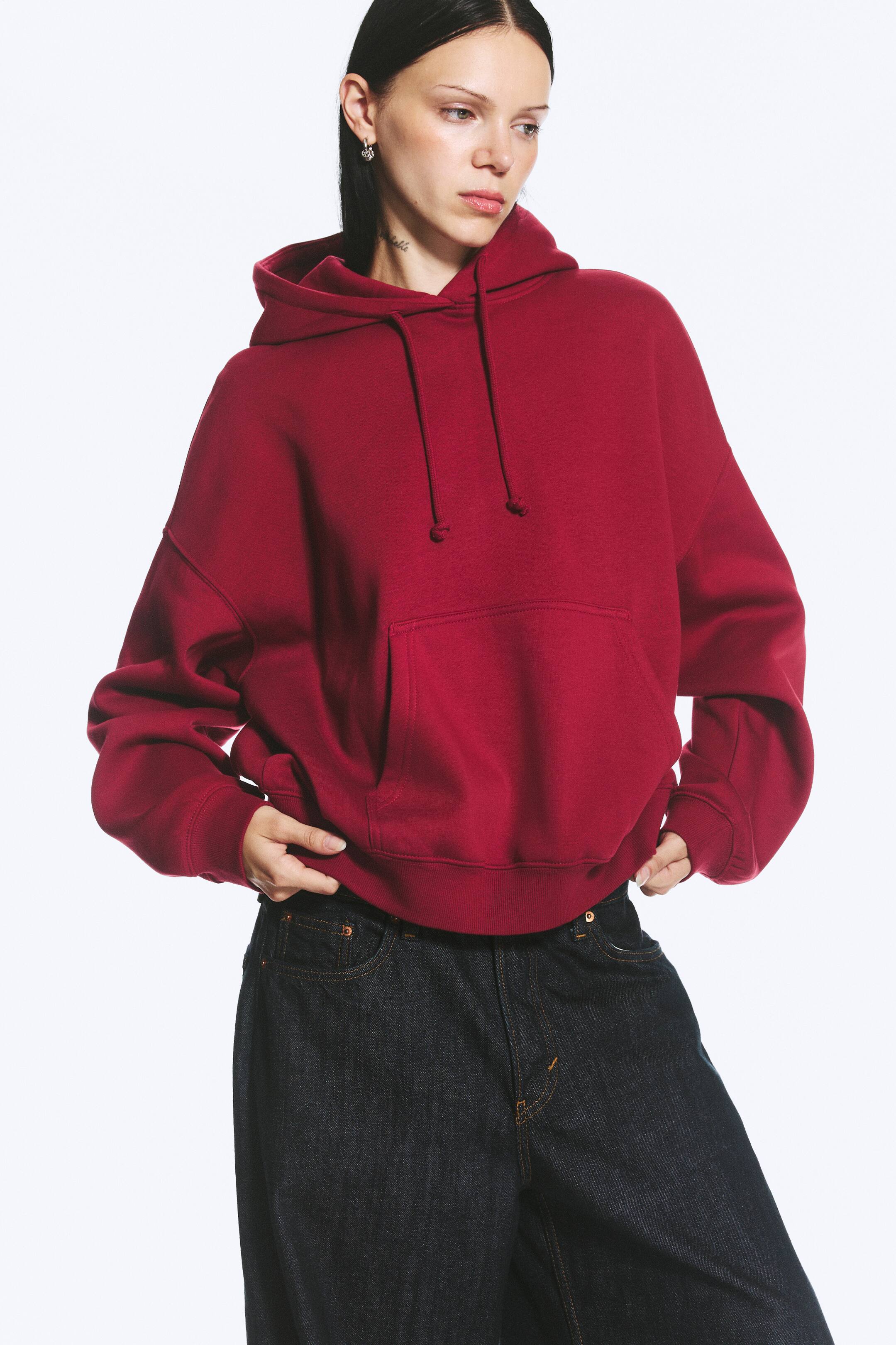 View larger image: A deep red regular-fit hoodie with a regular length and long sleeves is worn by the model. The hoodie features a roomy drawstring hood, dropped shoulders, a large kangaroo pocket, and ribbed cuffs and hem. The model is holding the ribbed hem with both hands.