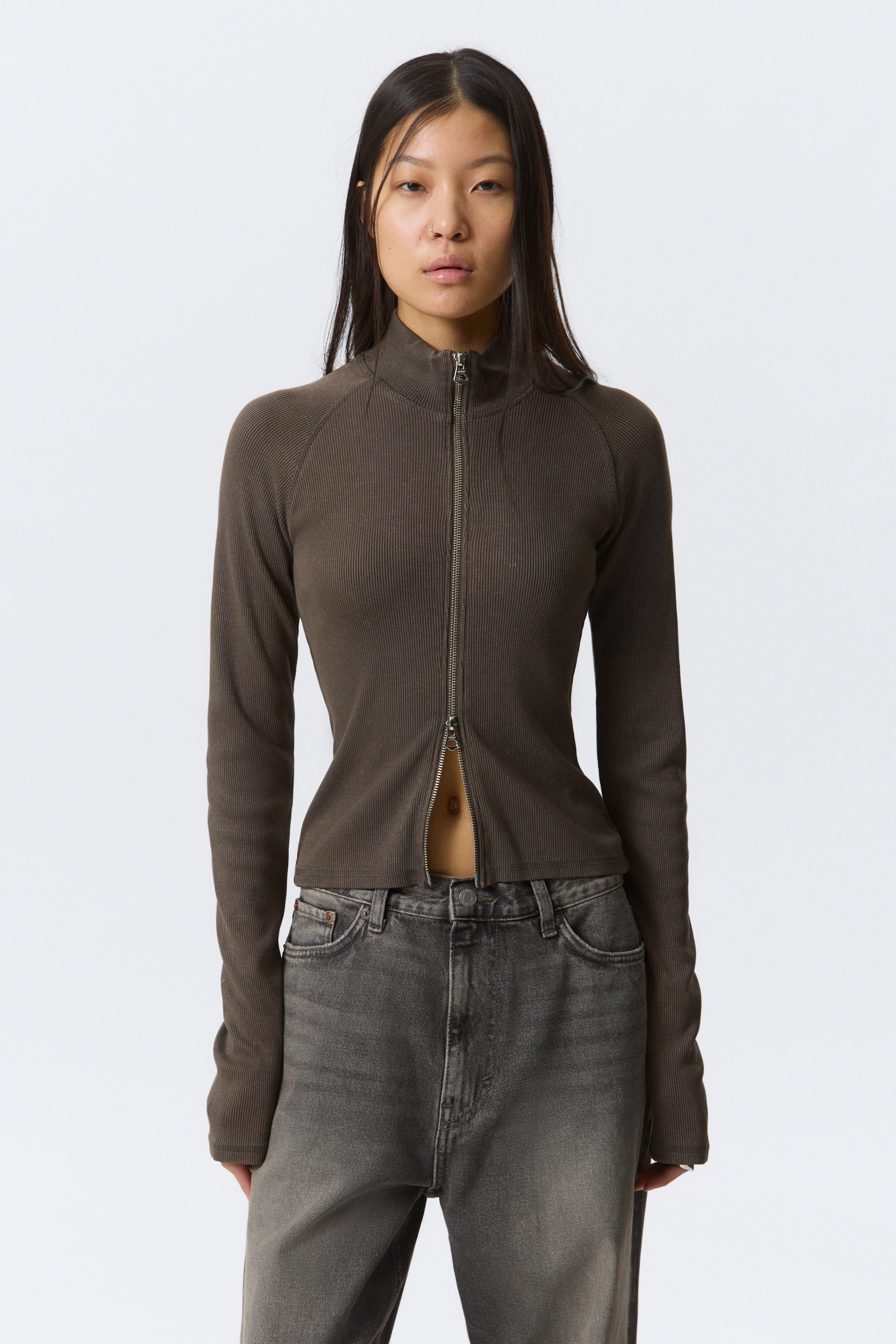 View larger image: A dark brown ribbed full-zip top is facing forward, featuring a mock neck collar and long raglan sleeves. A silver-toned zip runs down the front, partially unzipped from the bottom, showcasing a slim fit.