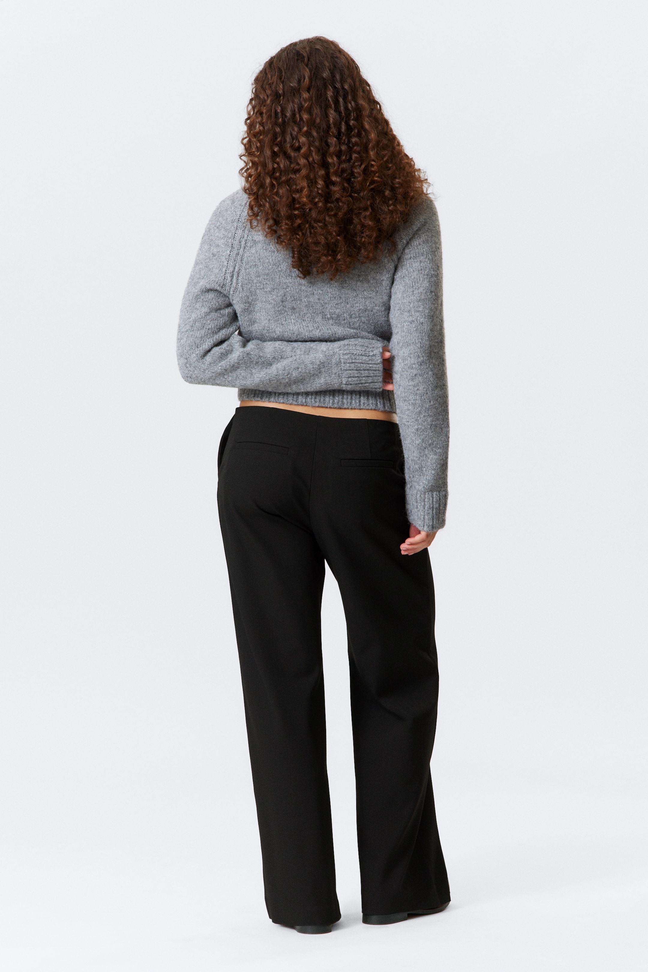 Grösseres Bild ansehen: Ein grauer Strickpullover mit Textur und schwarze Hosen mit weitem Bein werden von hinten präsentiert. Der kurze Pullover hat Raglanärmel, gerippte Bündchen und einen gerippten Saum, der einen schmalen Streifen des unteren Rückens über den hoch taillierten Hosen zeigt, die eine entspannte Passform und zwei rückseitige Paspeltaschen aufweisen.