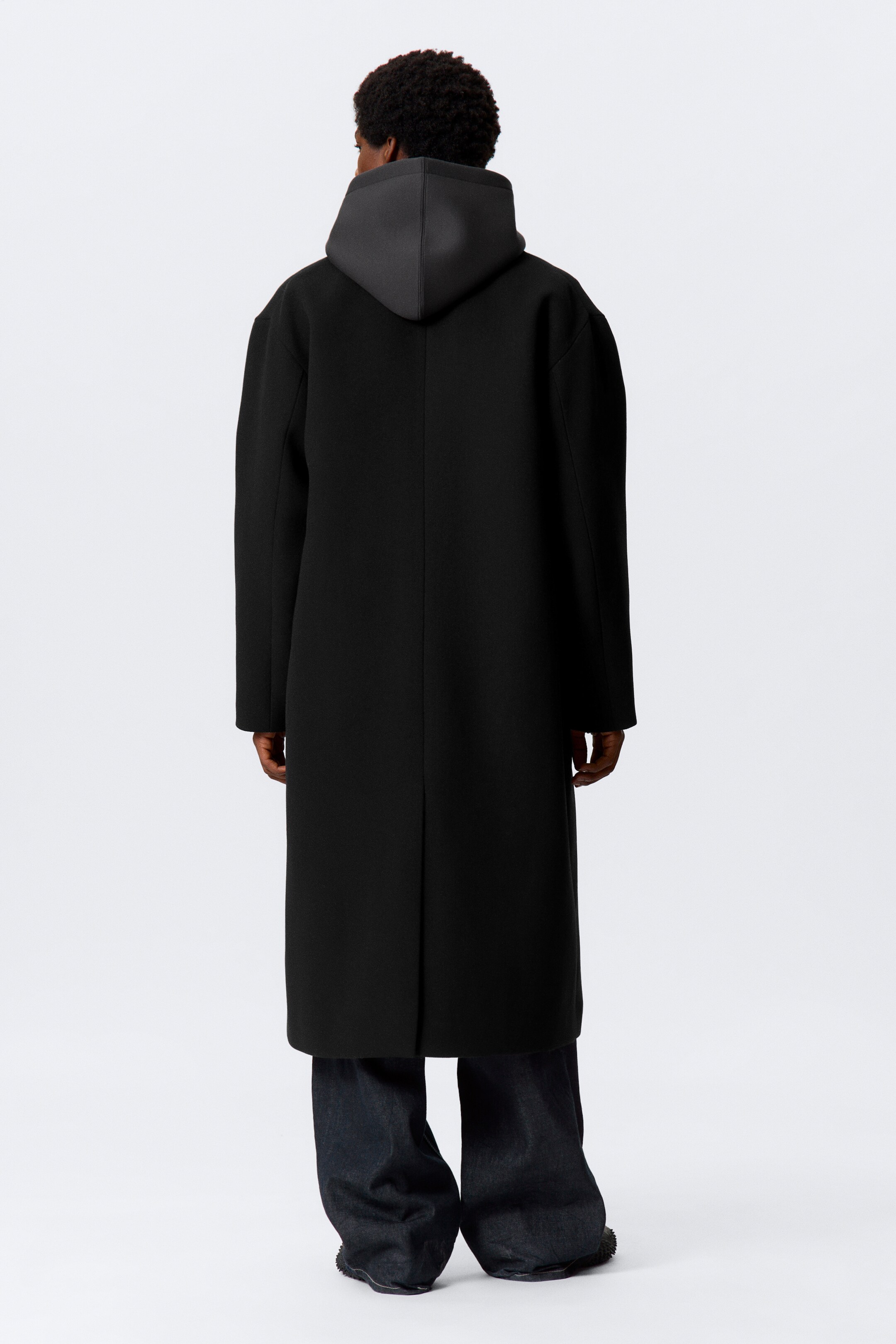 Größeres Bild ansehen: Ein schwarzer langer Mantel, der fast bis zu den Knöcheln reicht, wird von hinten betrachtet. Er präsentiert eine gerade, übergroße Silhouette mit breiten Schultern und einem langen Rückenschlitz, während ein dunkelgrauer Hoodie darunter geschichtet sichtbar ist und seine Kapuze auf den Schultern ruht.