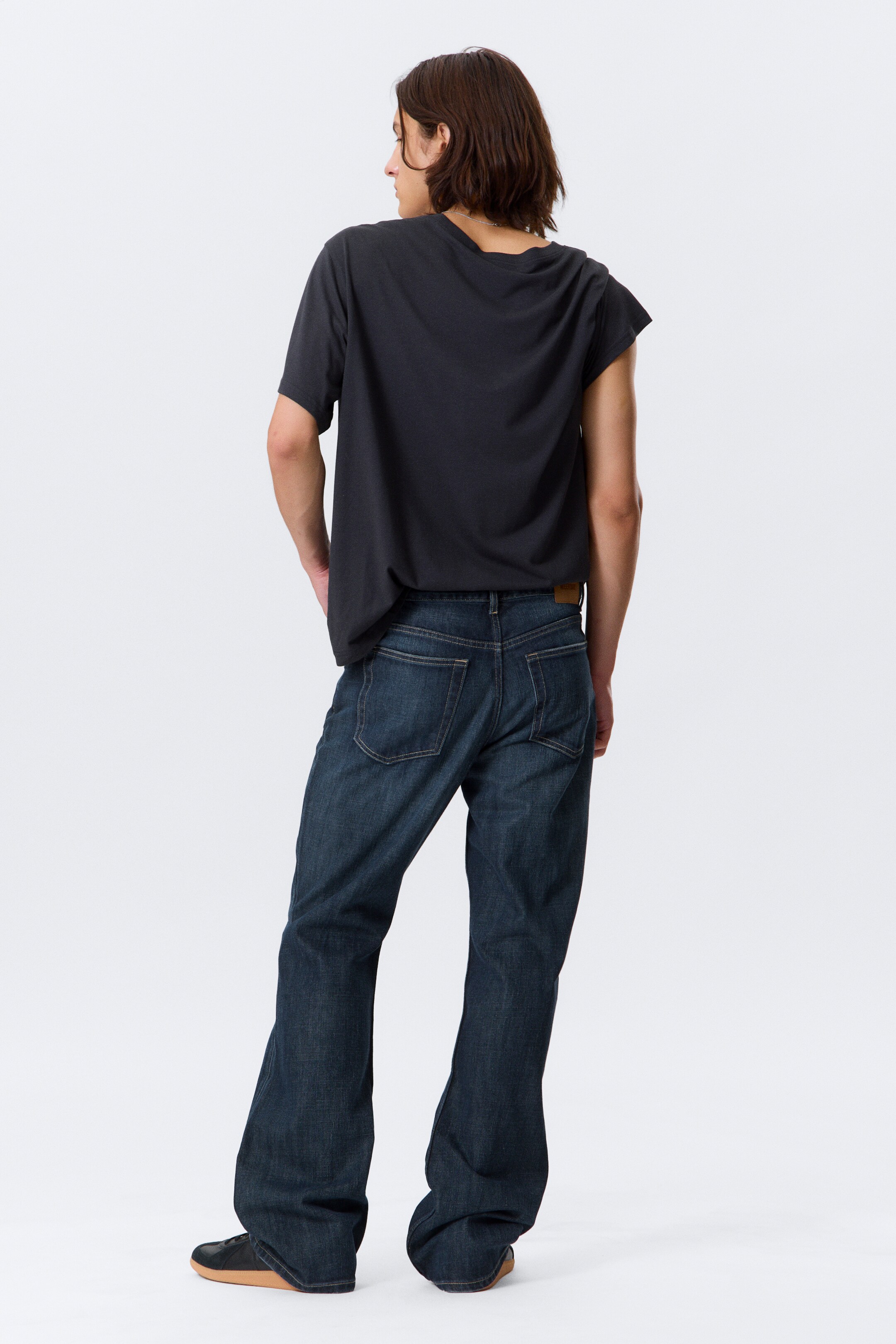 View larger image: A dark charcoal short-sleeved t-shirt and dark wash wide-leg jeans are being worn by a person angled away and slightly to their right. The relaxed t-shirt is draping loosely, while the jeans are showcasing visible back pockets and a brand patch on the waistband, with the fabric pooling around the ankles.