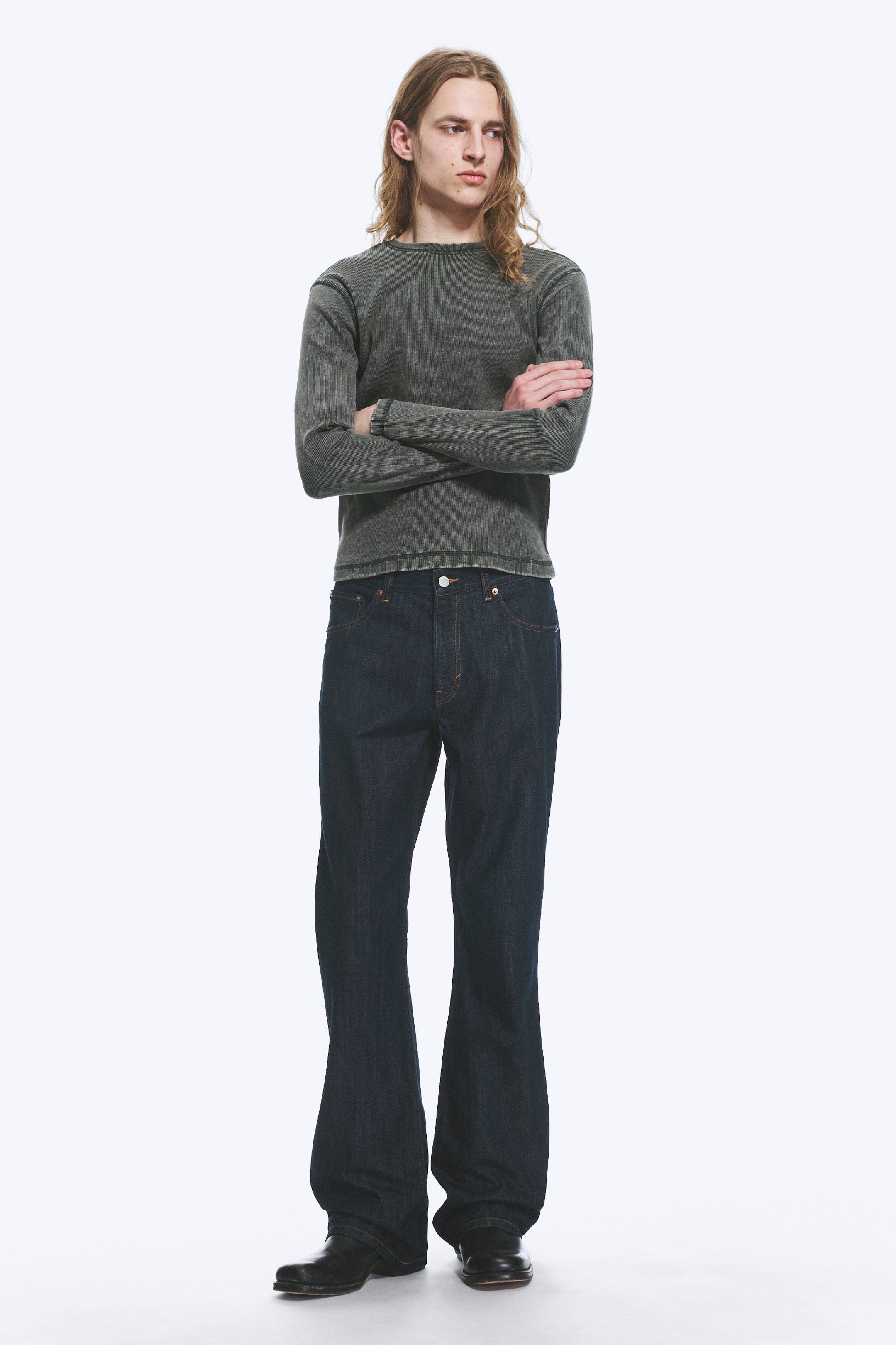 View larger image: A dark grey long-sleeved crewneck shirt, featuring prominent topstitched seams, is being worn. The shirt is paired with dark wash indigo bootcut jeans, showcasing a button fastening and contrast stitching, with their wide leg opening visibly flaring from the knee.