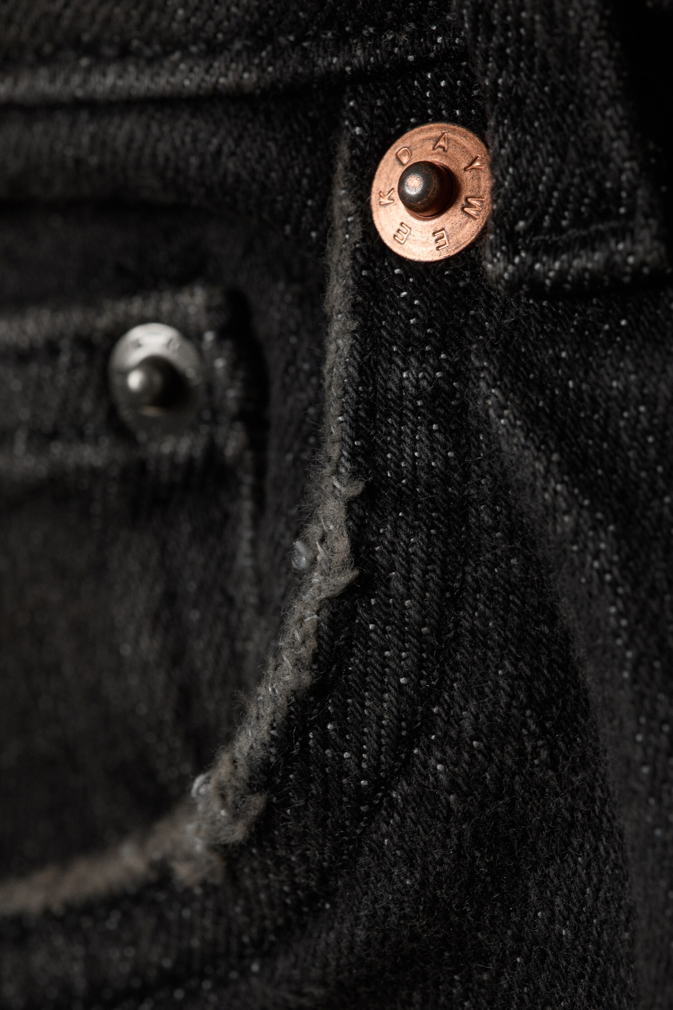 Grotere afbeelding bekijken: De voorkant van donkere denim jeans wordt in close-up getoond, met de focus op een kopertint knoop met gegraveerde letters en een wazige zilverkleurige klinknagel. De textuur van de stof heeft kleine witte vlekjes, en een gestikte naad met lichte rafeling is zichtbaar.