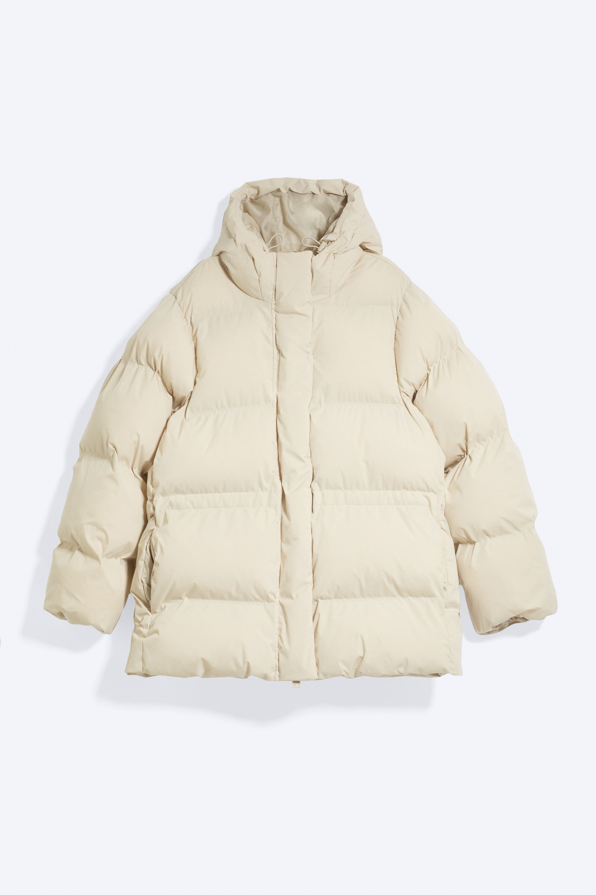 Das Model trÃ¤gt eine cremefarbene, taillierte Pufferjacke mit horizontaler Steppung, Kapuze und geraffter Taille. Die Jacke ist teilweise offen gestylt, Ã¼ber einem dunklen Oberteil getragen und mit lÃ¤ssig verwaschenen, weit geschnittenen blauen Jeans kombiniert.