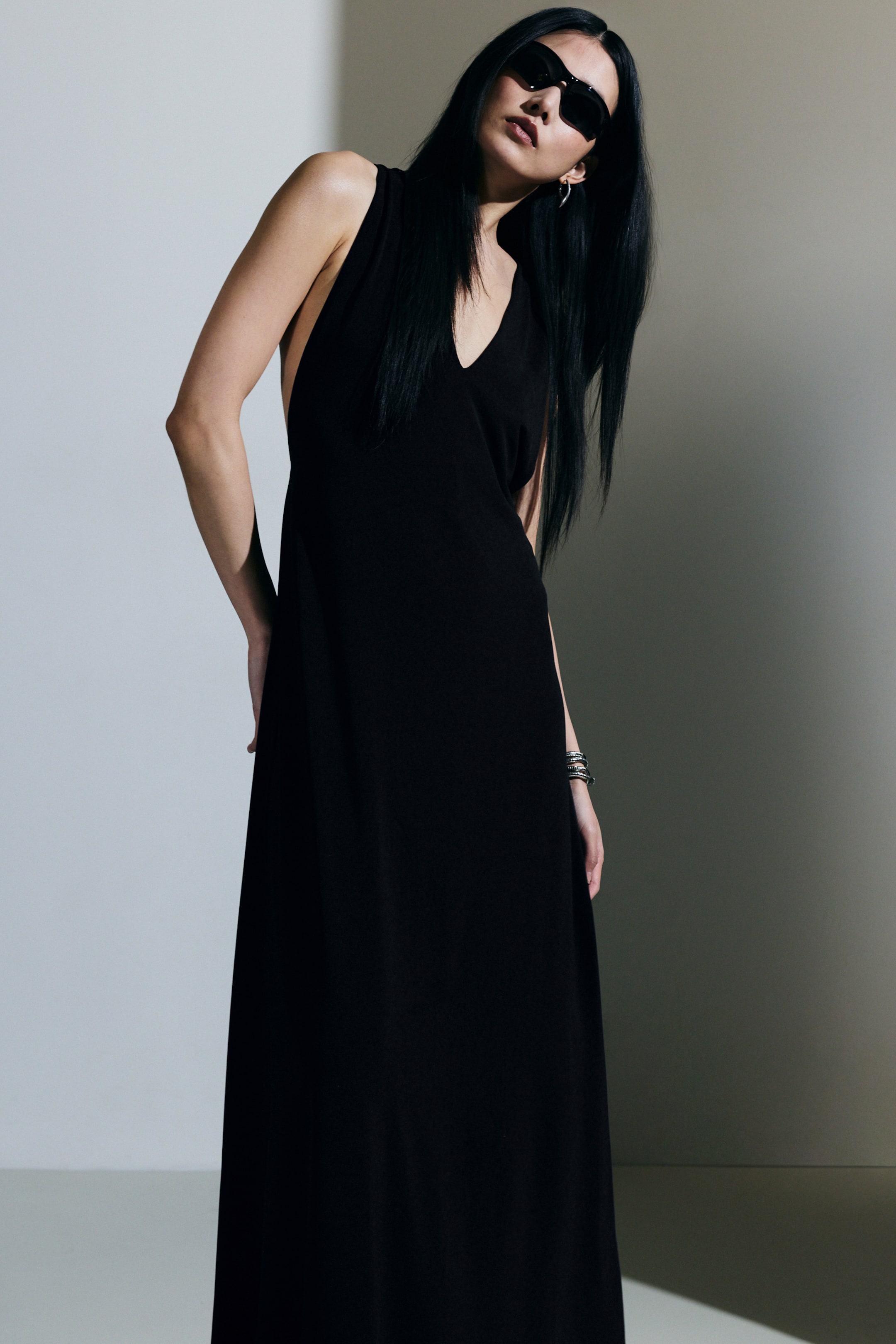 Größeres Bild anzeigen: Gerafftes Maxikleid. Dieses schwarze Maxikleid zeichnet sich durch einen V-Ausschnitt und tiefe Armausschnitte aus. Das Modell trägt das Kleid, das eine locker sitzende Silhouette hat.