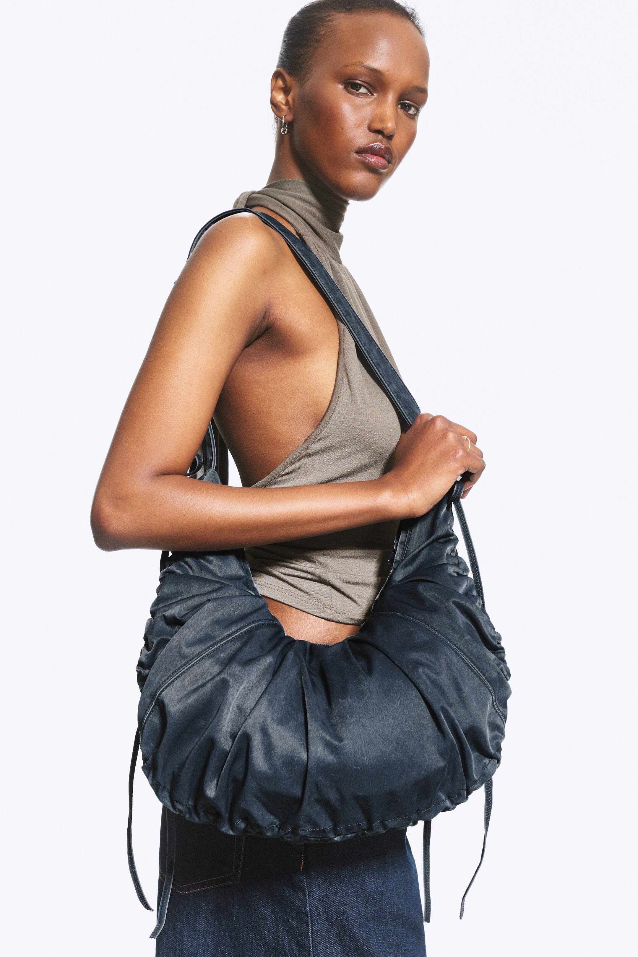 Vezi imaginea mai mare: O geantă de umăr tip crossbody de culoare închisă, cu detalii încrețite și șnururi, este purtată de model. Ea o poartă pe un umăr, ținând cureaua.