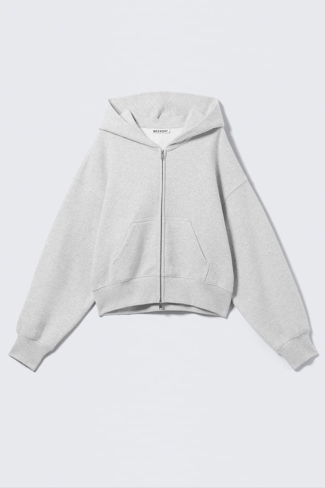 Ein hellgrauer Zip-up-Hoodie wird von VORN gezeigt, gefertigt aus weicher Baumwolle mit boxy-Silhouette und Cropped-Länge. Er verfügt über eine locker sitzende Kapuze, einen zentralen Front-Reissverschluss, eine Kängurutasche sowie gerippte Bündchen an Ärmeln und Saum. - 1
