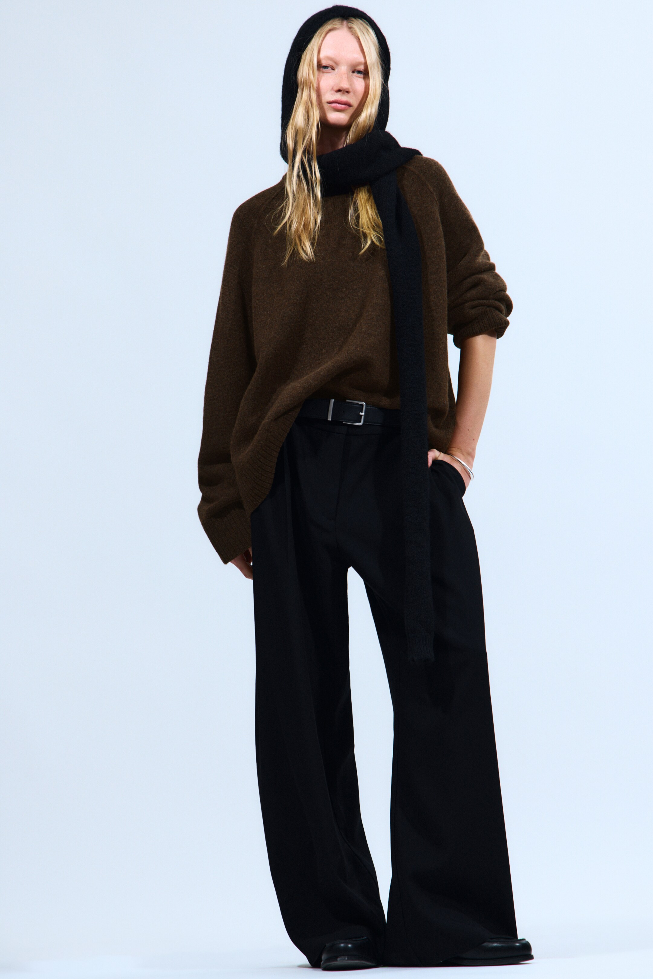 Agrandir l'image: Un pull en maille marron surdimensionné avec des manches longues et des poignets côtelés est porté, stylisé avec un pantalon noir ample à jambes larges avec des plis à l'avant et une ceinture noire avec une boucle argentée. Une capuche en maille noire couvre la tête, et une écharpe en maille noire est drapée autour du cou, complétant le look en couches.