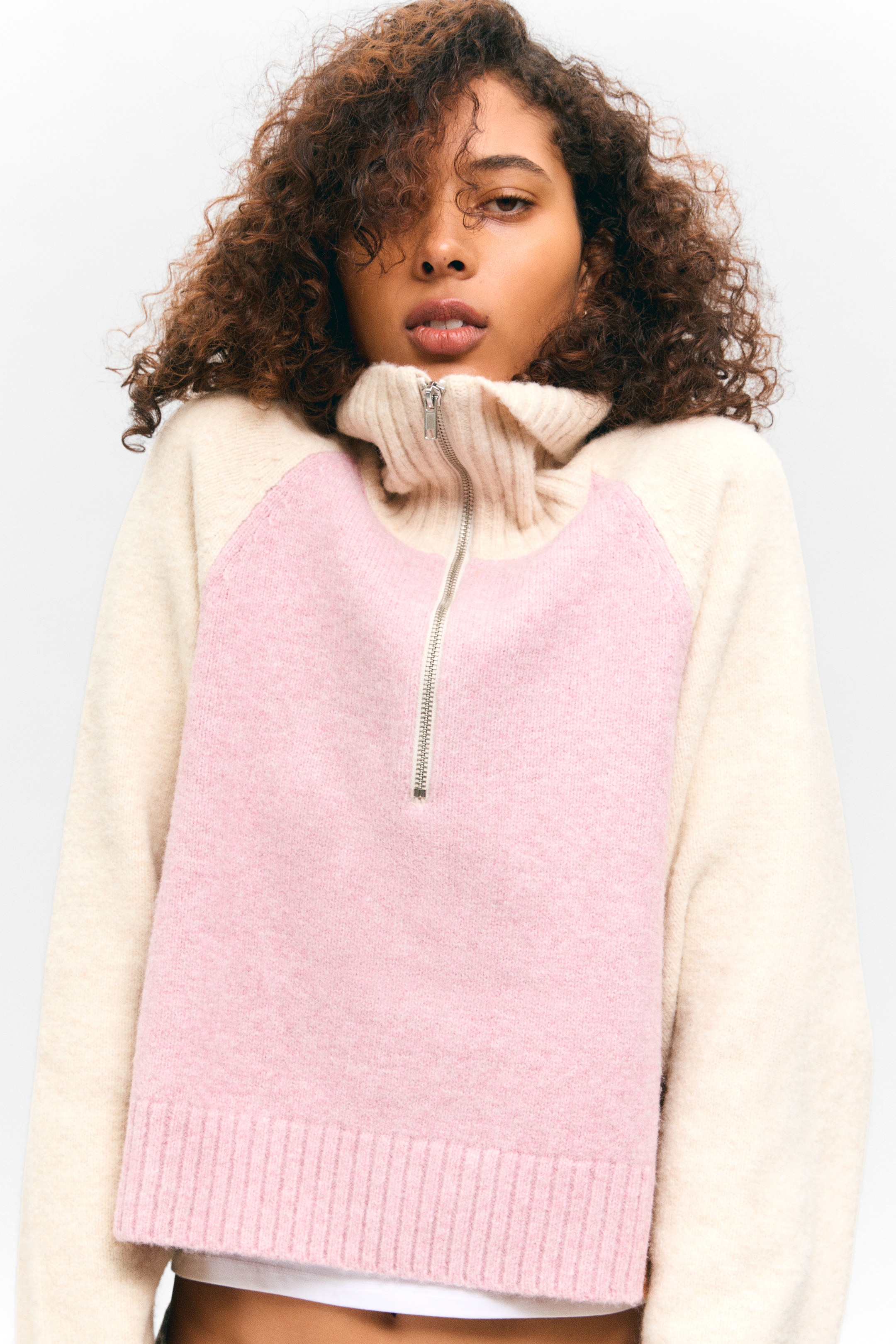 Grösseres Bild ansehen: Ein flauschiger Color-Block-Strickpullover präsentiert eine pinkfarbene Vorderseite mit cremefarbenen Raglanärmeln. Er ist mit einem gerippten, cremefarbenen hohen Kragen und einem silberfarbenen Halbreißverschluss, der sich an der Vorderseite nach unten erstreckt, gestaltet, und der untere Saum ist ebenfalls gerippt und pink.