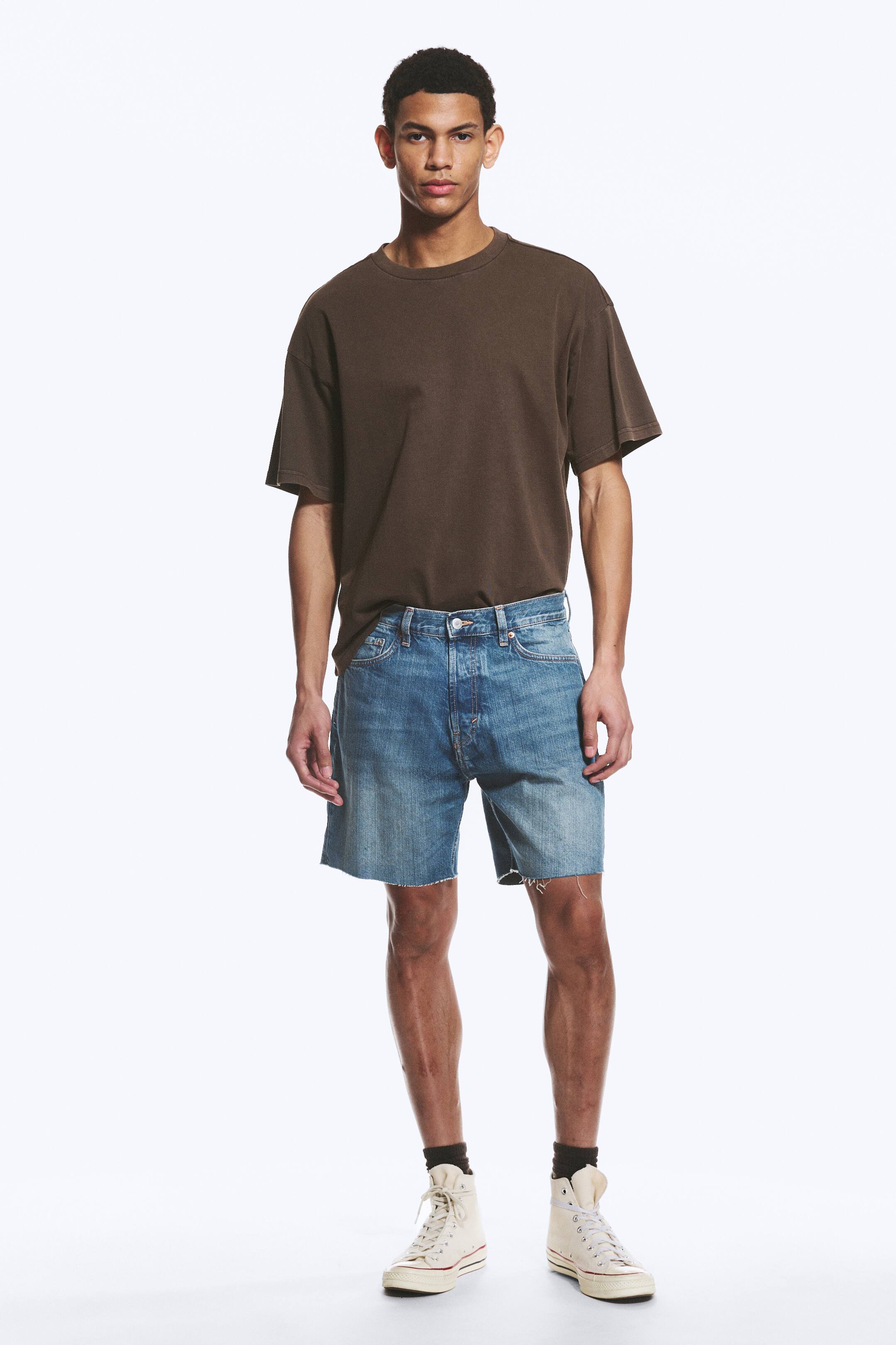 Visa större bild: En brun oversized t-shirt med rund hals bärs tillsammans med ljusblå denimshorts med knappgylf och råa, slitna fållar som sträcker sig till mitten av låret. Outfiten kompletteras med bruna strumpor och krämfärgade höga snörskor.