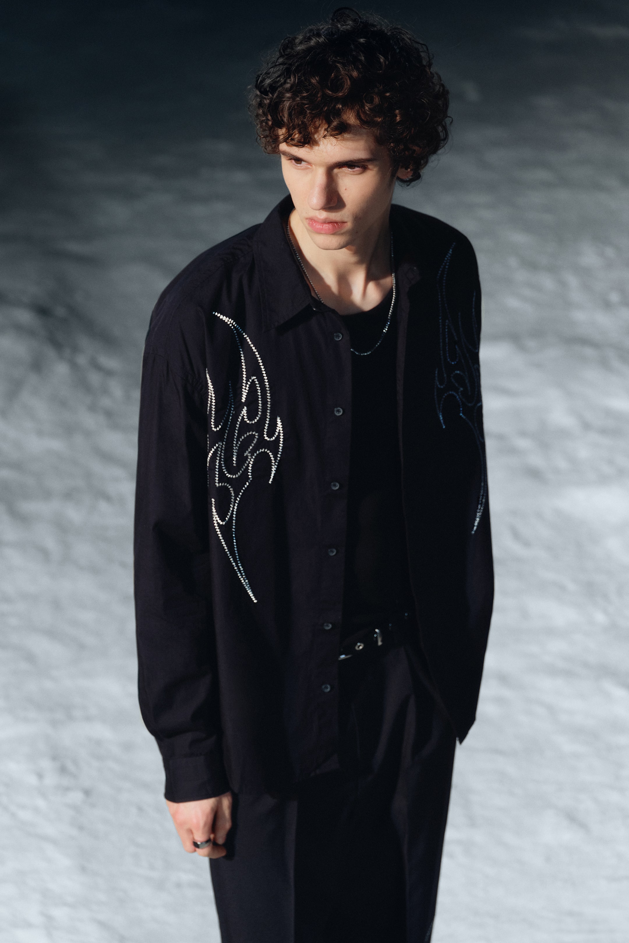 Vis større bilde: En svart skjorte med knapper presenteres fra en litt forhøyet, venstre forfra vinkel. Den har lange ermer, en spiss krage og sølvfargede, rhinestone-pyntede flammemotiver som dekorerer frontpanelene. Skjorten bæres åpen over en svart undertrøye, som avslører et tynt sølvfarget kjedehalsbånd, og er kombinert med svarte bukser.