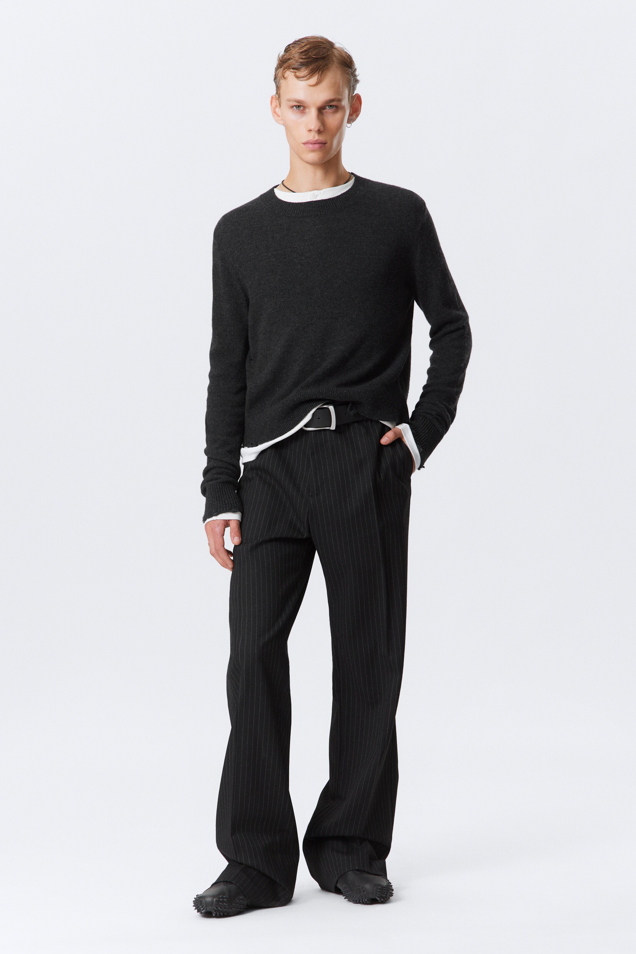 View larger image: A dark gray knitted sweater with a slim fit and shrunken silhouette is being worn by the model. It features a crew neckline and subtle distressing at the hems and cuffs, with a white layer visible underneath.
