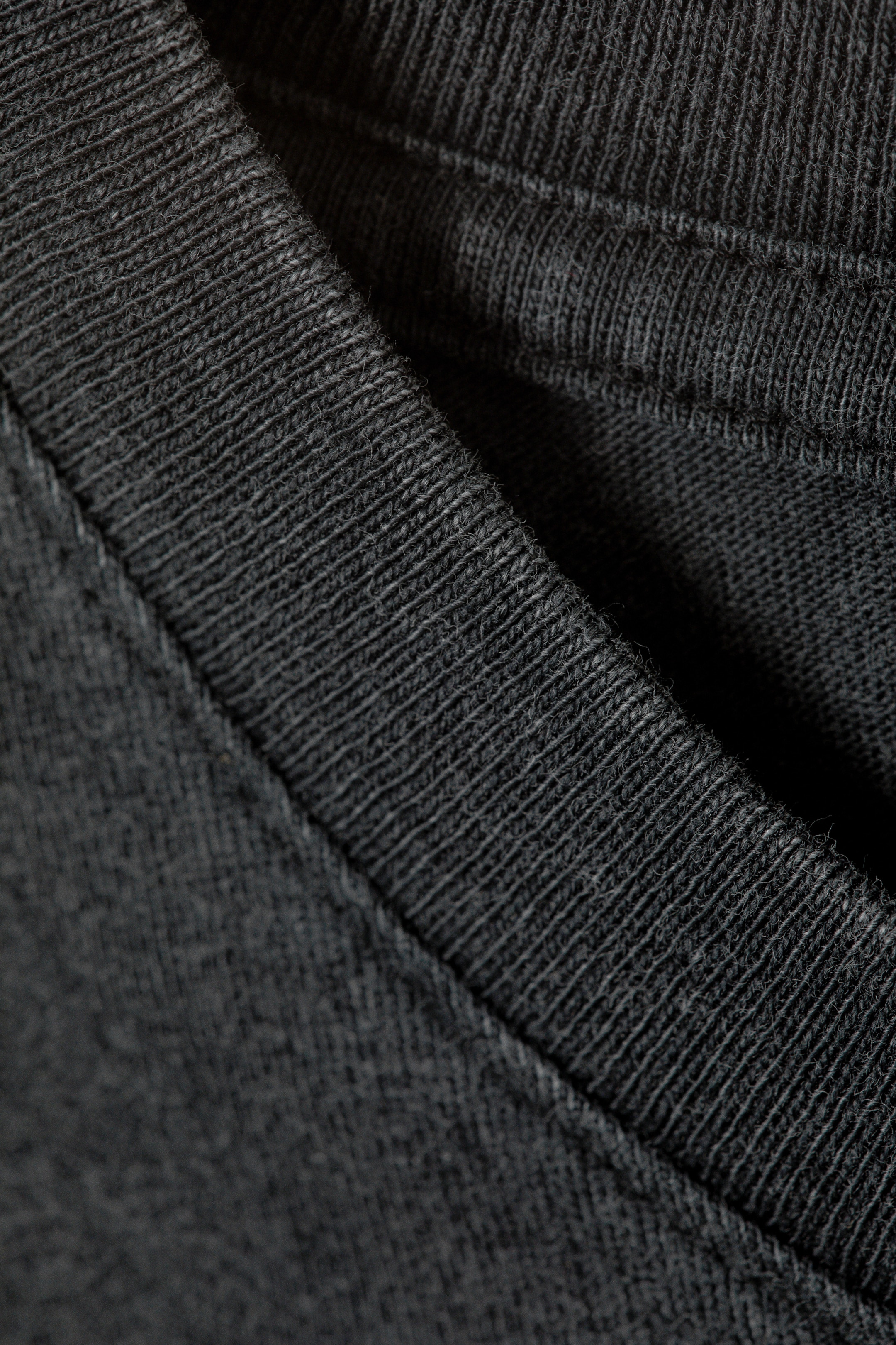 View larger image: The dark grey crewneck T-shirt's neckline is featuring a ribbed band with twin-needle topstitching running parallel on the main jersey knit fabric. Clean chain stitching is visible at the neckline seam, connecting the ribbed band to the T-shirt's body.