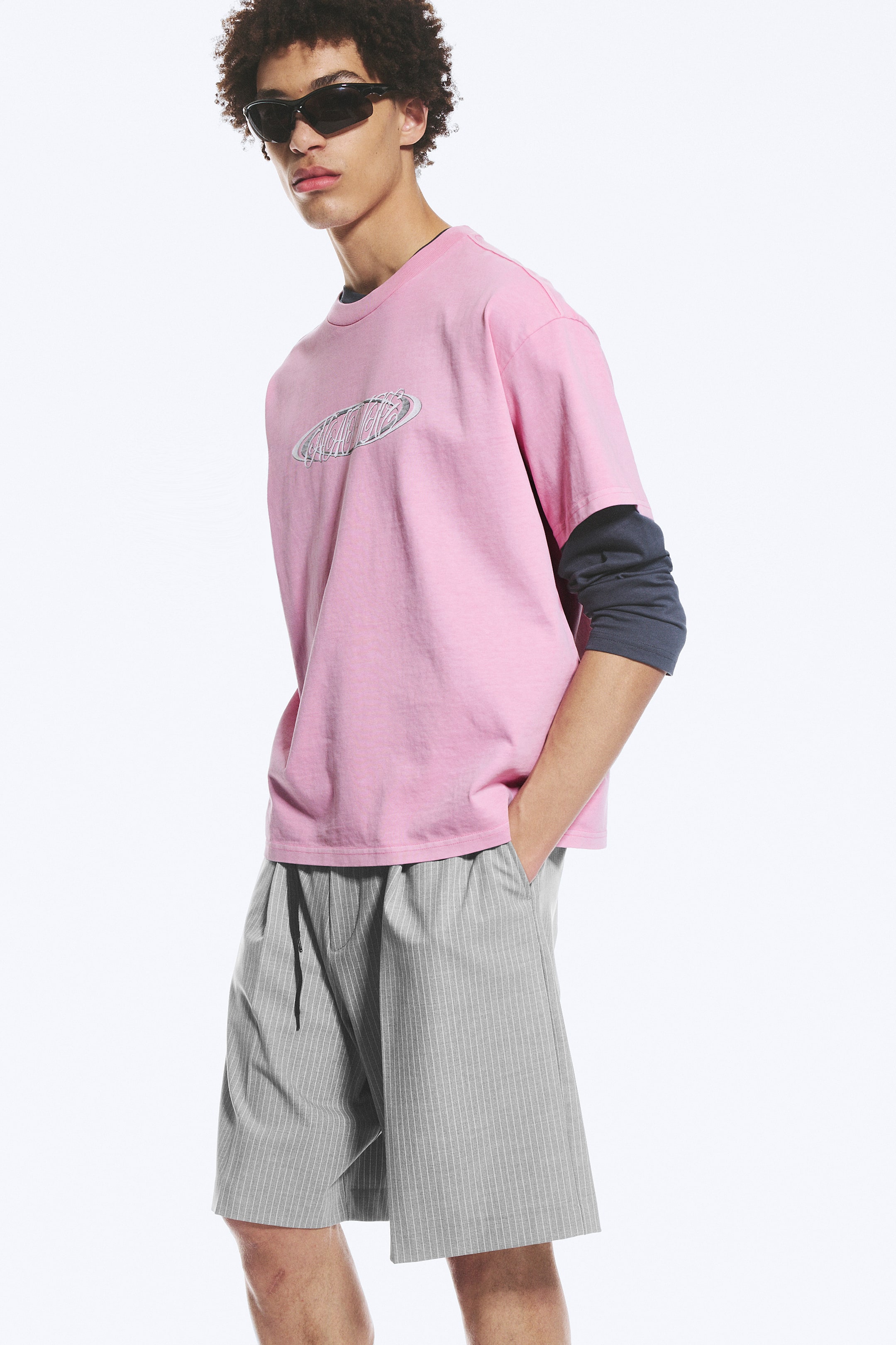 Visualizza l'immagine ingrandita: Una t-shirt rosa a maniche corte presenta un grafico ovale in tonalità argento con testo wireframe centrato sul petto. Mostra una vestibilità rilassata ed è indossata sopra una maglia a maniche lunghe scura, con le maniche dello strato inferiore che si estendono oltre i polsini della t-shirt.