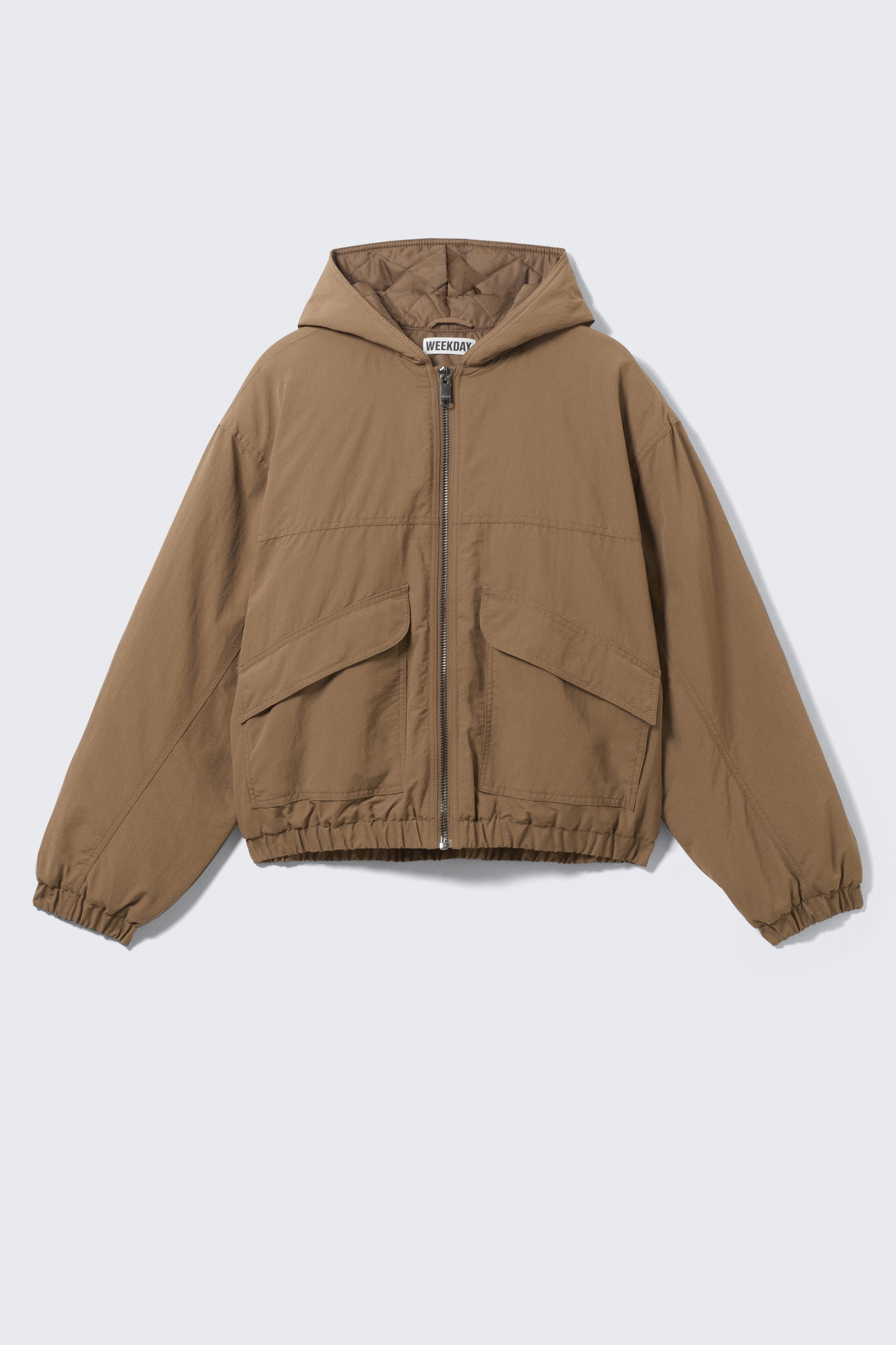 Größeres Bild anzeigen: Eine braune Nylon-Bomberjacke mit Kapuze wird von vorne gezeigt, sie hat eine entspannte Passform und eine geräumige Kapuze mit sichtbarem Steppfutter innen. Sie zeigt auch einen robusten zentralen Reißverschluss, zwei Klettverschluss-Patten-Taschen und elastische Bündchen und einen elastischen Saum.