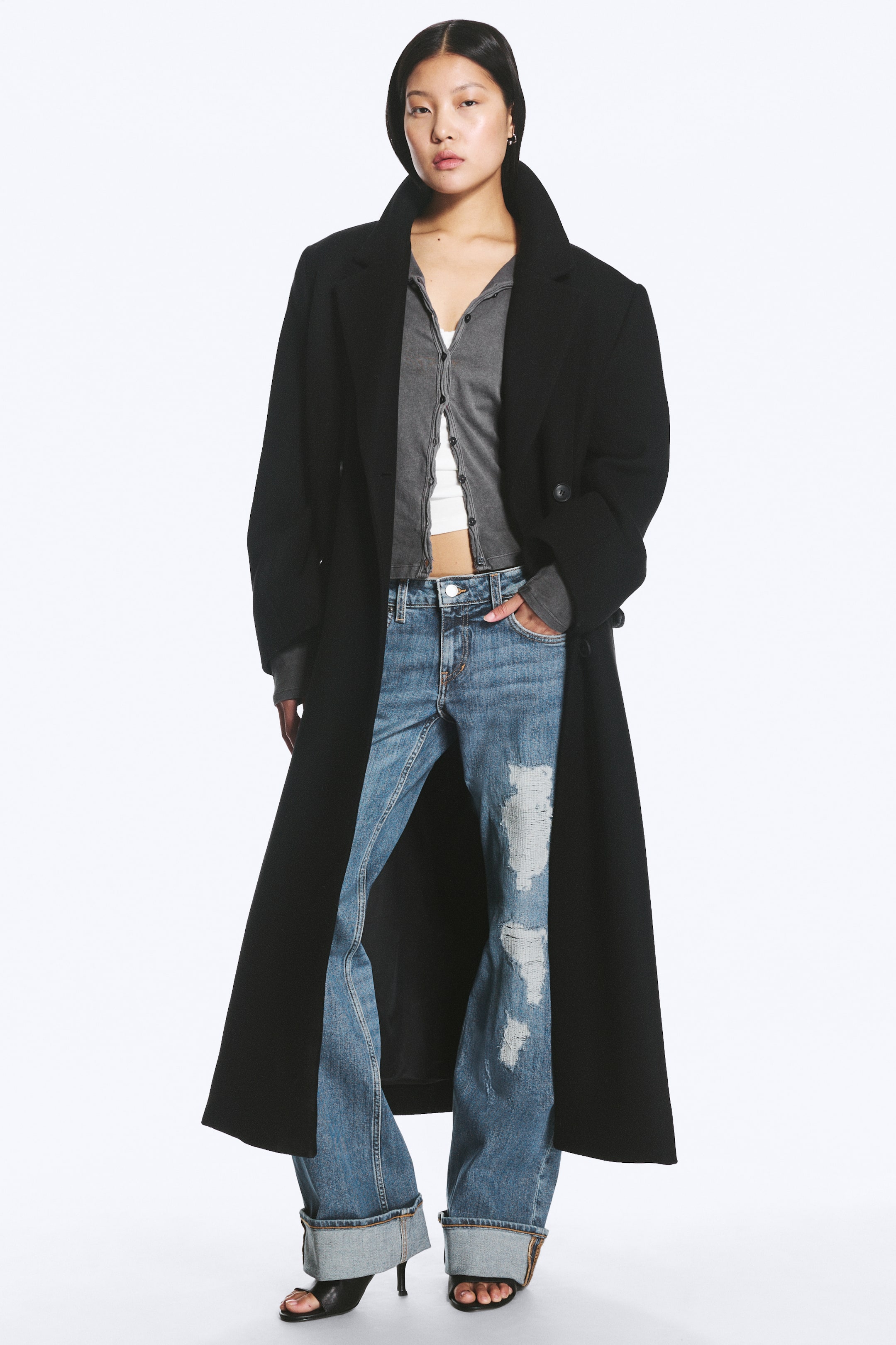 Zobacz większy obraz: Płaszcz LOGAN DOUBLE-BREASTED WOOL-BLEND jest noszony przez modelkę rozpięty, co eksponuje jego czarną, wydłużoną sylwetkę, szerokie ramiona i klapy z wycięciem. Został wystylizowany na szary kardigan i biały crop top.