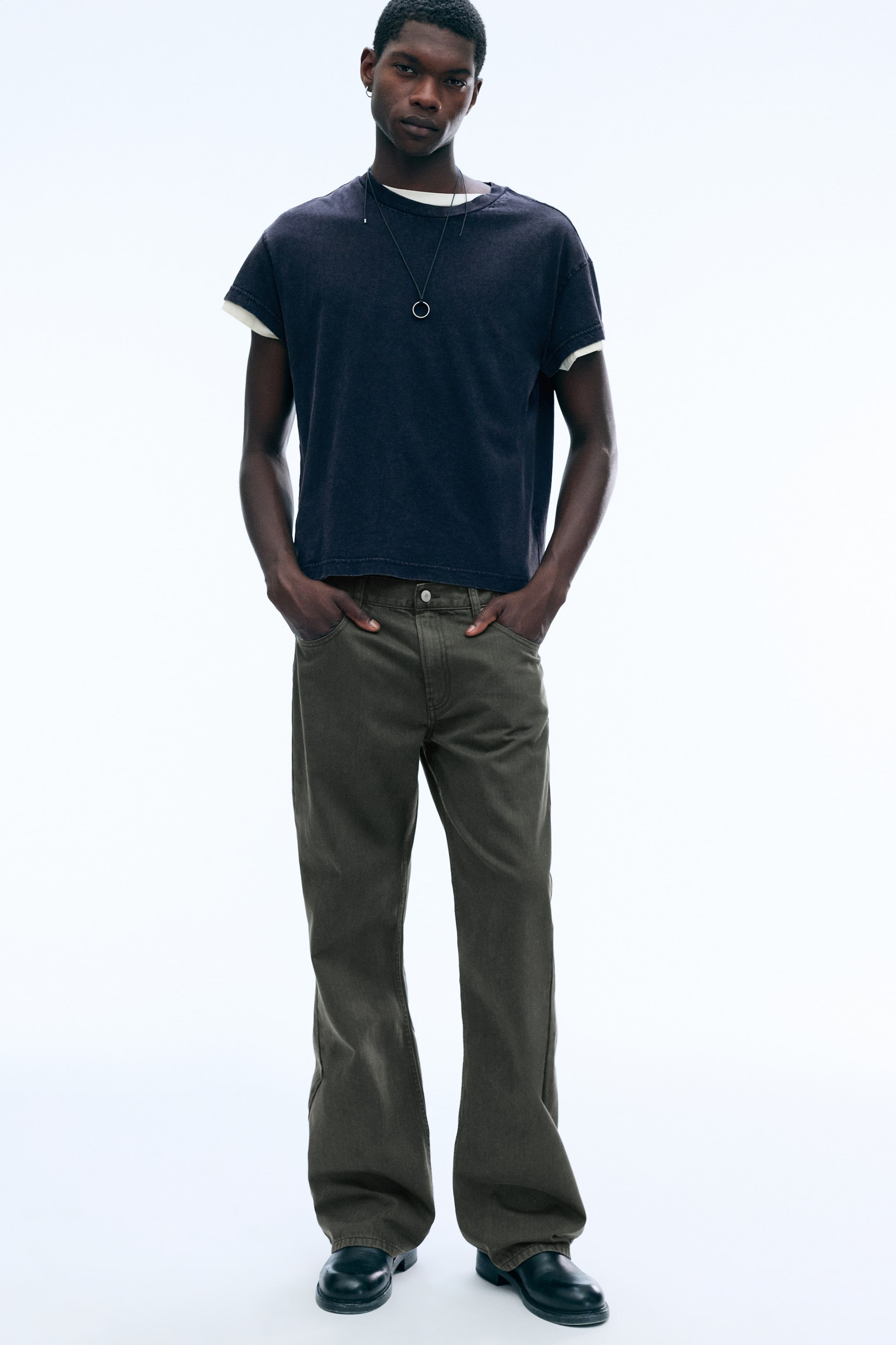 Visa större bild: Mercury bootcut-byxor med fem fickor i mörkt olivgrönt bärs av modellen, som står med händerna vilande i framfickorna. Byxorna har en klassisk femficksdesign och en lätt utsvängd benvidd, stylade här med en lager-på-lager-inspirerad mörk t-shirt och svarta stövlar.