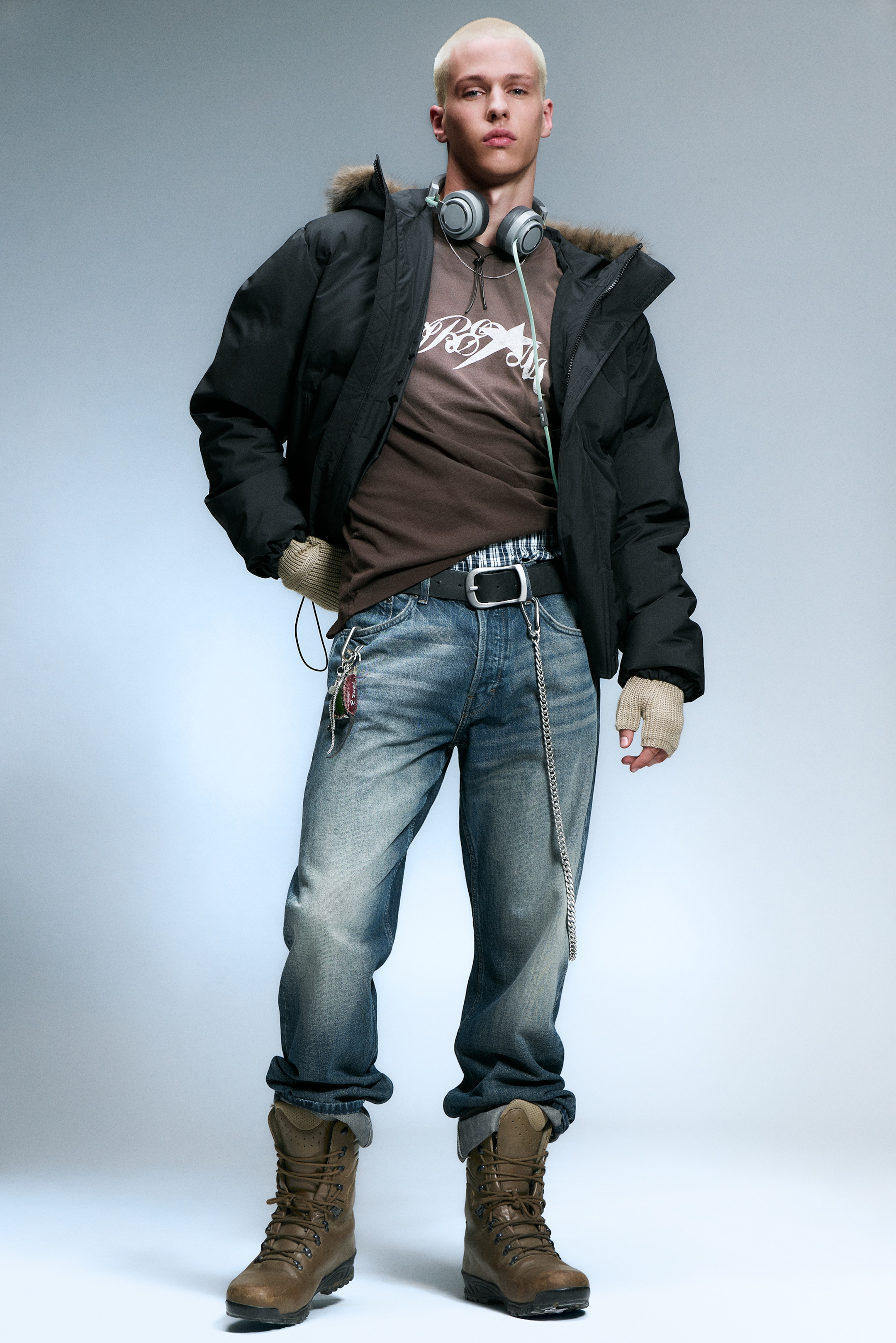 Größeres Bild ansehen: Eine Frontalansicht eines Outfits zeigt eine schwarze Daunenjacke mit einem pelzbesetzten Kapuze, die über einem braunen Grafikt-T-Shirt und verblassten blauen Jeans liegt, die an den Knöcheln umgeschlagen sind. Braune Schnürstiefel vervollständigen die untere Hälfte, während ein schwarzer Gürtel mit einer silbernen Schnalle, eine silberne Kette mit Schlüsseln, graue Kopfhörer und hellfarbige fingerlose Handschuhe den Look accessorieren.