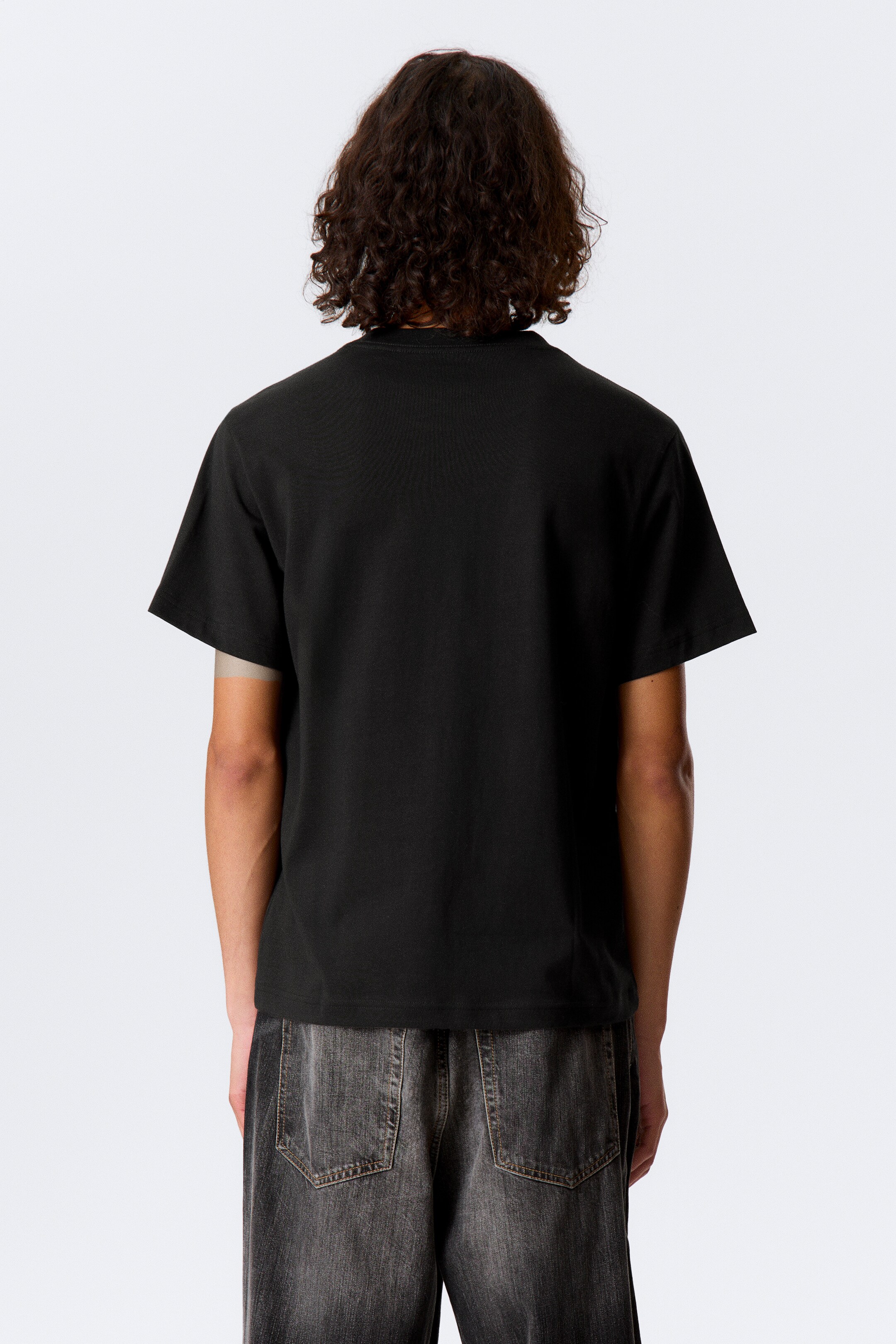 Ver imagem maior: Uma t-shirt preta de mangas curtas está a ser vista de costas. Apresenta uma gola redonda e uma bainha reta, oferecendo um ajuste descontraído sobre jeans de lavagem escura.