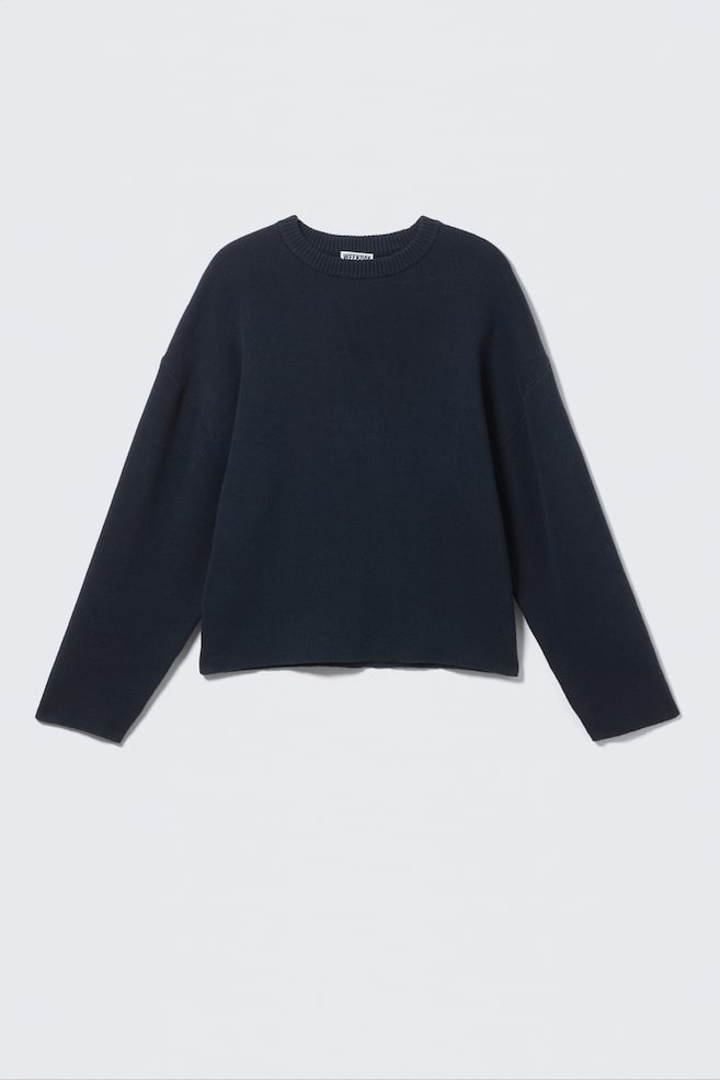 En mørkeblå, boxy-fit sweater fremstillet af blød bomuldsstrik præsenteres forfra. Den har en klassisk rund halsudskæring med en ribbet kant og et 'WEEKDAY'-mærke, nedfaldne skuldre og brede ærmer. Rene, glatte sømme afslutter manchetterne og bunden, hvilket skaber et sømløst udseende. - 1