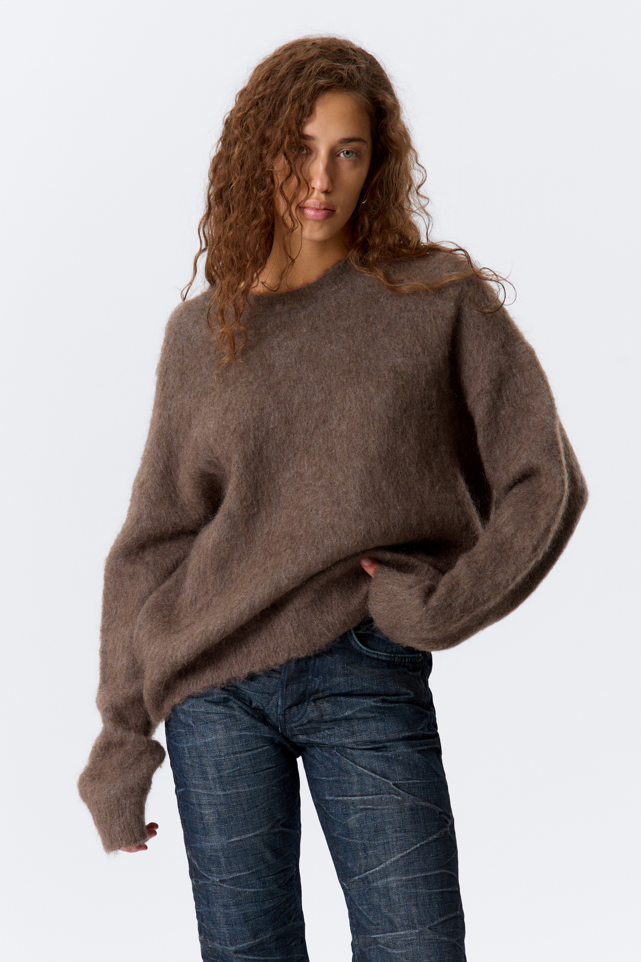 Größeres Bild ansehen: Ein brauner, haarig strukturierter Pullover wird aus einer frontalen, leicht angewinkelten Perspektive präsentiert, mit einem Rundhalsausschnitt und langen, lockeren Ärmeln, deren Bündchen bis über die Hände reichen, und weist einen übergroßen Schnitt auf.