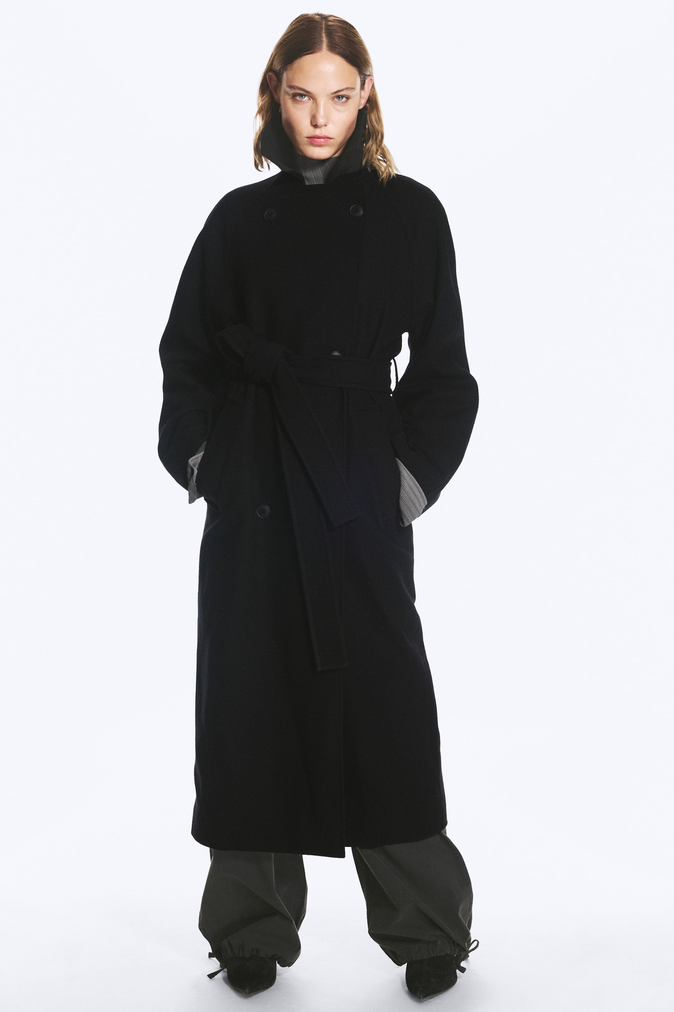 Visualizza immagine più grande: Il modello indossa un cappotto lungo nero oversize doppiopetto, con le mani infilate nelle tasche laterali. Il cappotto è abbinato a una cintura in tessuto annodata in vita, con colletto alto sovrapposto a un dolcevita grigio a coste e pantaloni scuri.