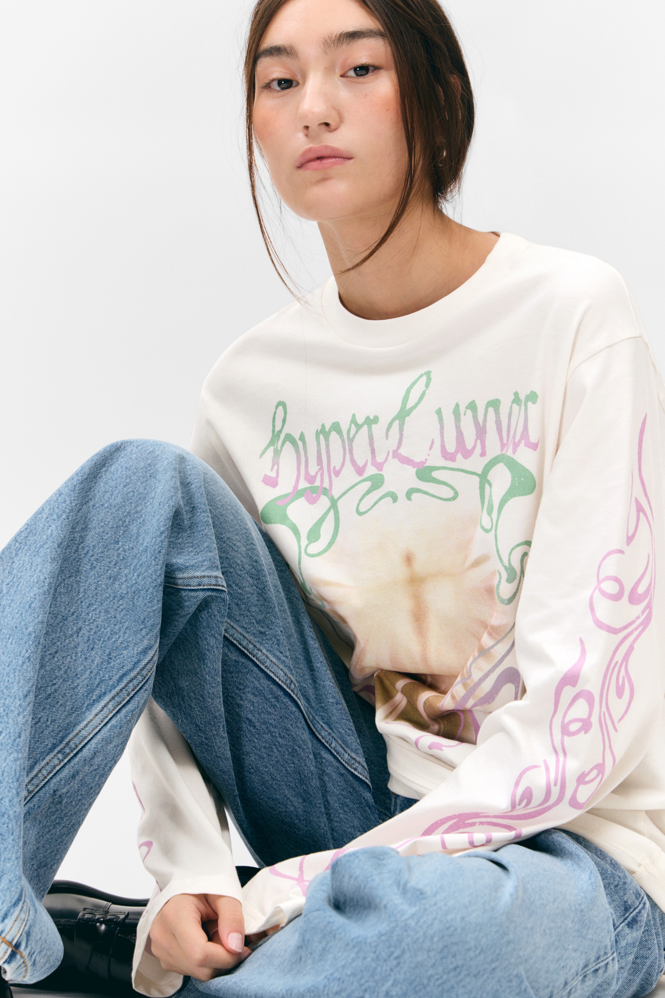 View larger image: An off-white, long-sleeved crew neck top is featuring "hyper Luna" text in green and purple script over a faded, multi-toned circular graphic on the front. Swirling purple patterns are extending down both sleeves, and the top is draping loosely over blue jeans.