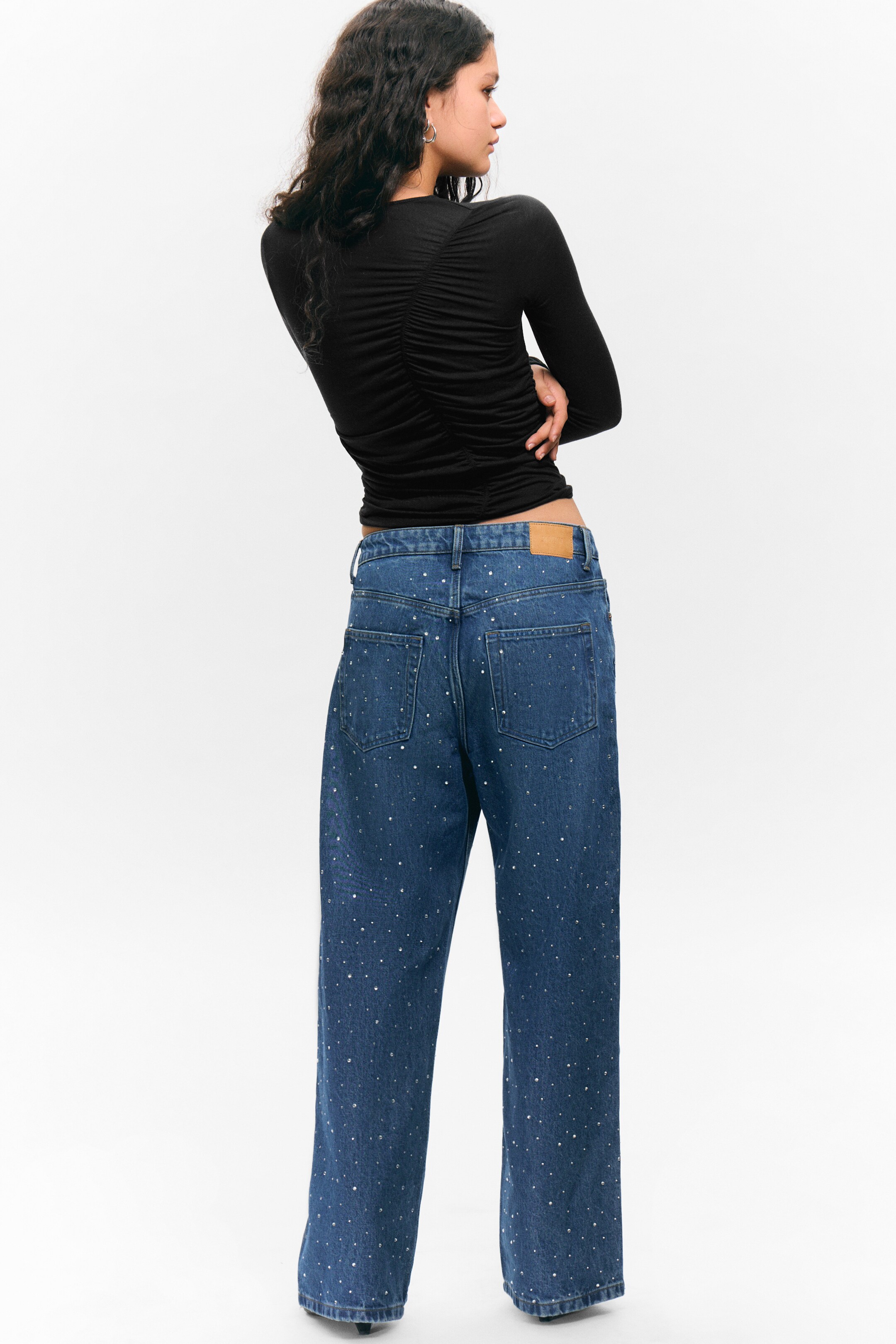 Visualizza immagine più grande: I jeans in denim blu sono presentati dal retro, leggermente inclinati verso destra. Presentano un taglio diritto, due tasche posteriori applicate, una toppa sulla cintura di colore marrone chiaro e sono completamente impreziositi da piccoli borchie scintillanti.