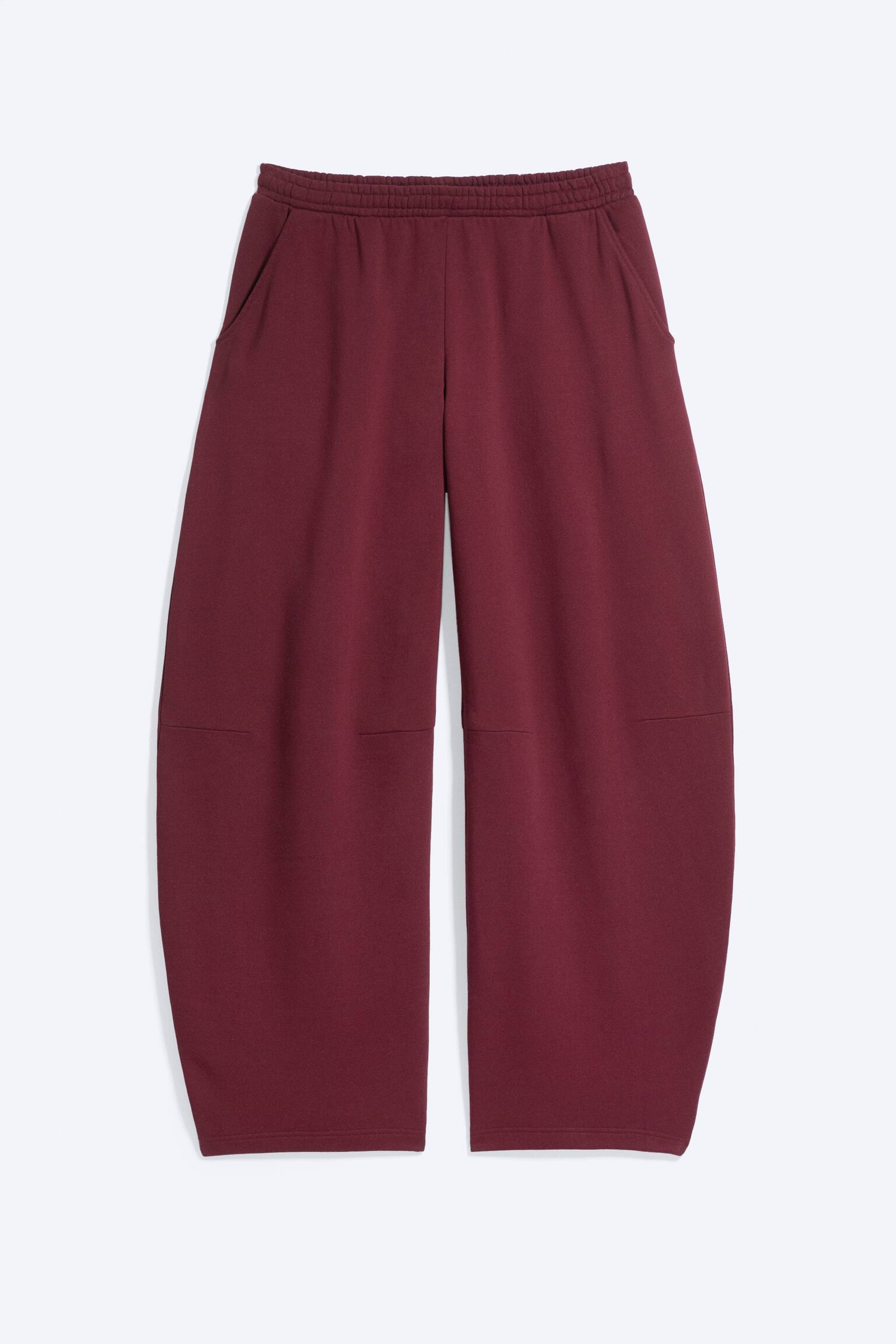 Un paio di pantaloni della tuta bordeaux dalla vestibilitÃ  morbida Ã¨ mostrato frontalmente, con vita elasticizzata e tasche laterali oblique. I pantaloni presentano una silhouette ampia a gamba barrel, che si allarga verso l'esterno, con cuciture di sagomatura orizzontali all'altezza delle ginocchia.