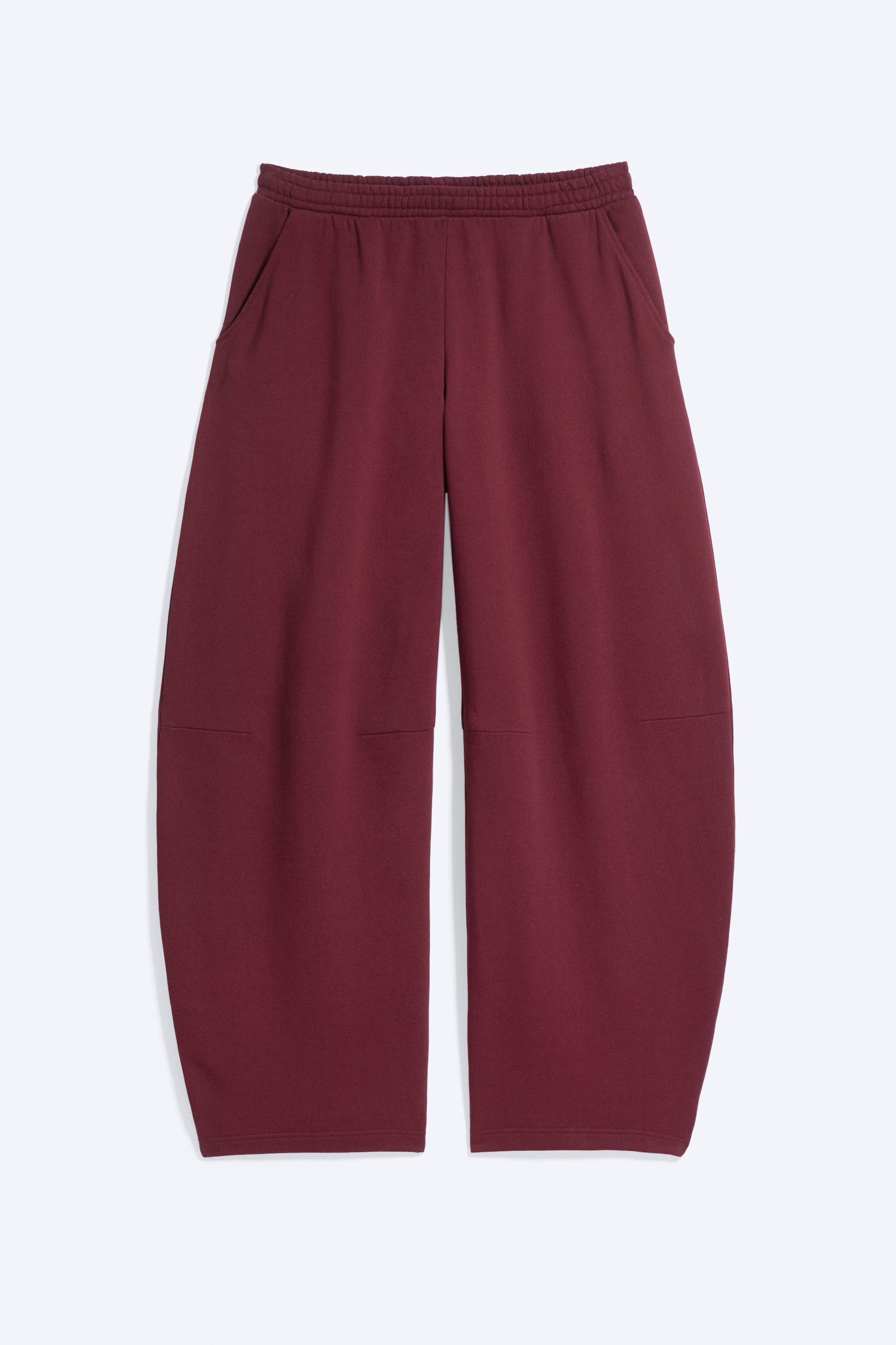 Größeres Bild ansehen: Ein Paar burgunderfarbene, locker geschnittene Sweatpants ist von vorne zu sehen und verfügt über einen elastischen Bund sowie schräge Seitentaschen. Die Hose zeigt eine weite, tonnenförmige Beinsilhouette, die sich nach außen wölbt, mit waagerechten formgebenden Nähten über den Knien.