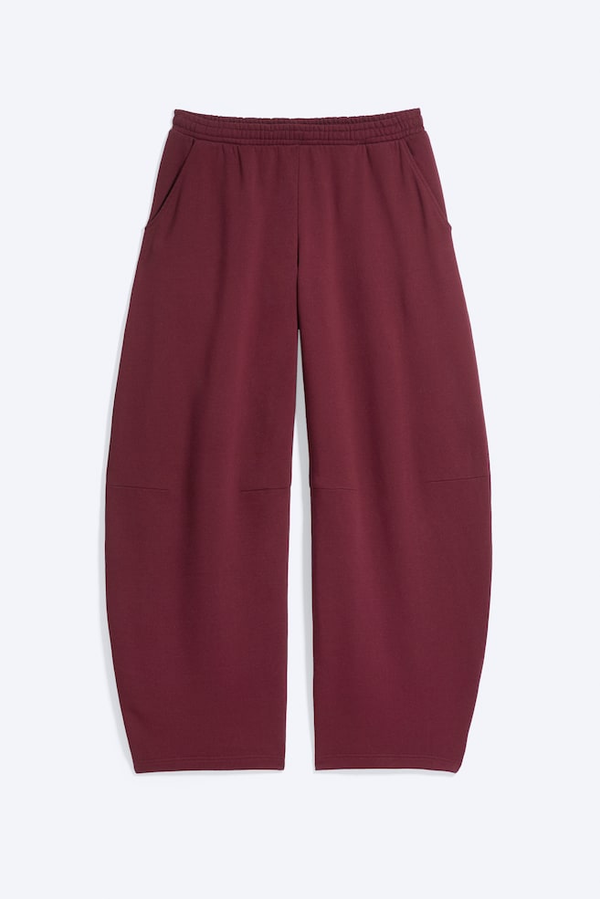 Se muestra un par de pantalones de chándal holgados color burdeos, vistos de frente, con cintura elástica y bolsillos laterales inclinados. Los pantalones presentan una silueta ancha de pernera de forma barril, que se curva hacia afuera, con costuras horizontales de marcado a la altura de las rodillas. - 1